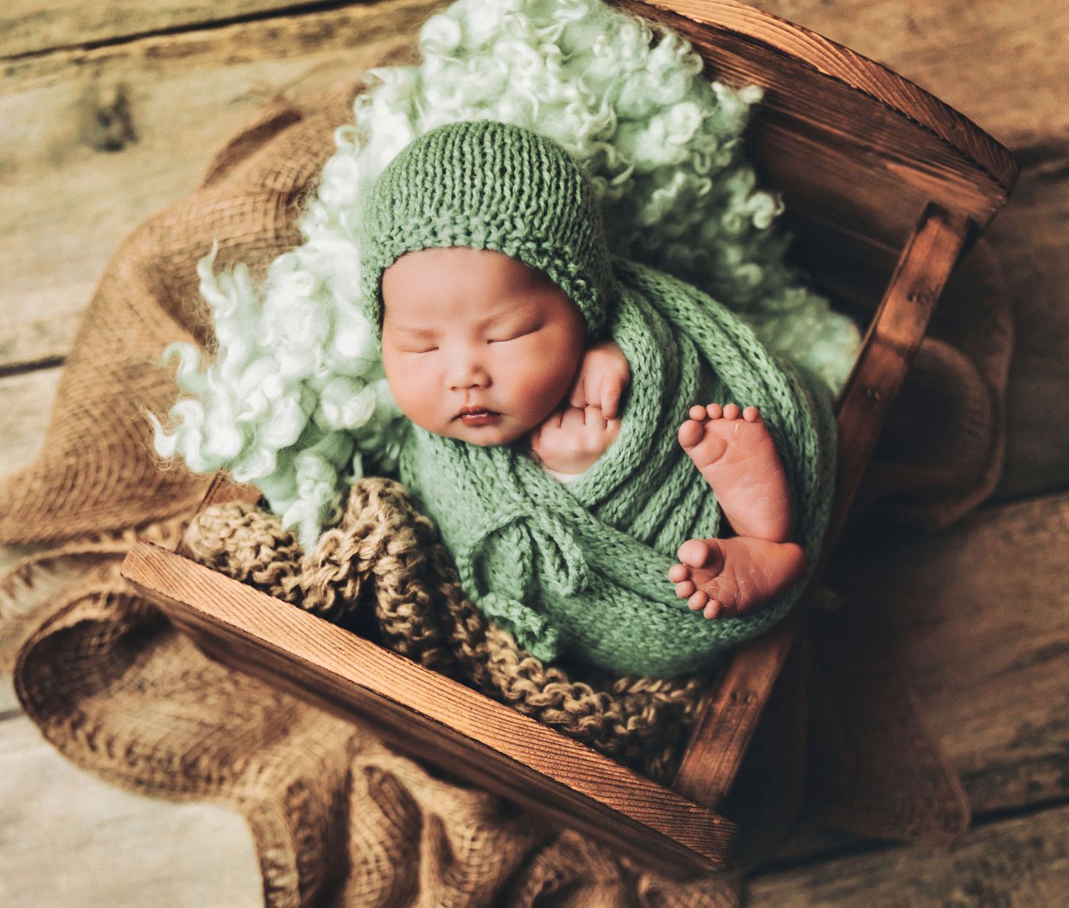 Newborn baby sleeping in wooden box wearing green knit hat and wrap with floral accents.