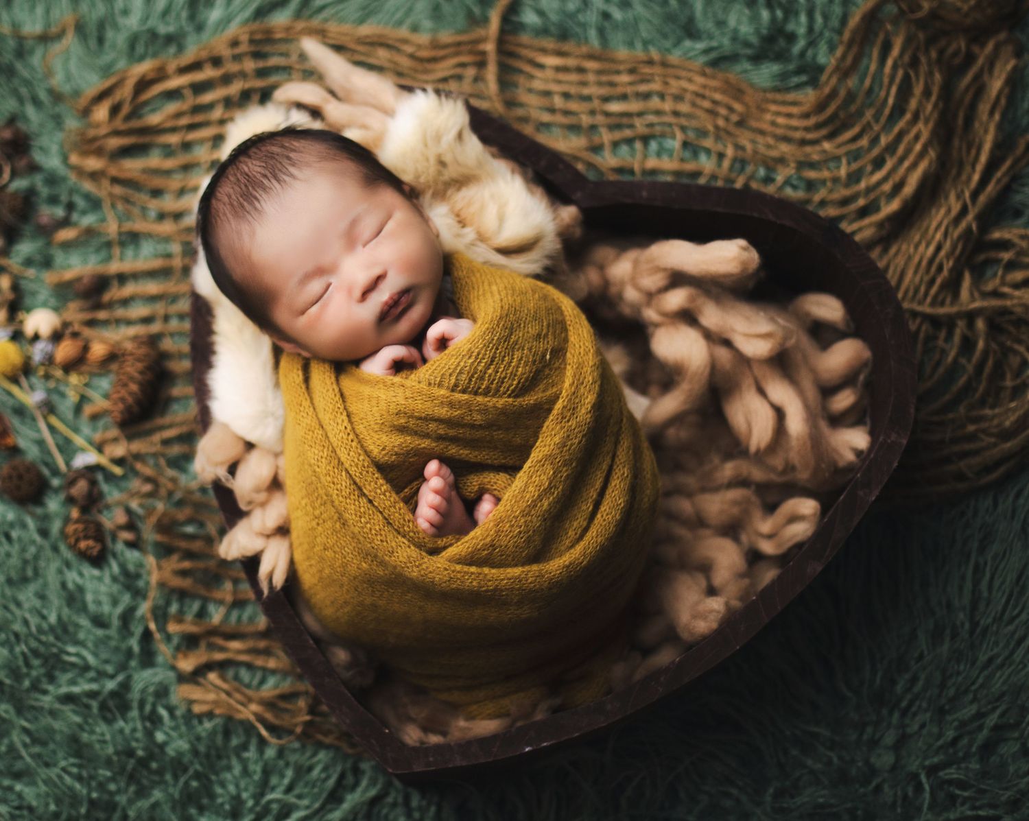 Newborn baby swaddled in mustard yellow wrap sleeping peacefully in a heart-shaped wooden bowl with soft textures.