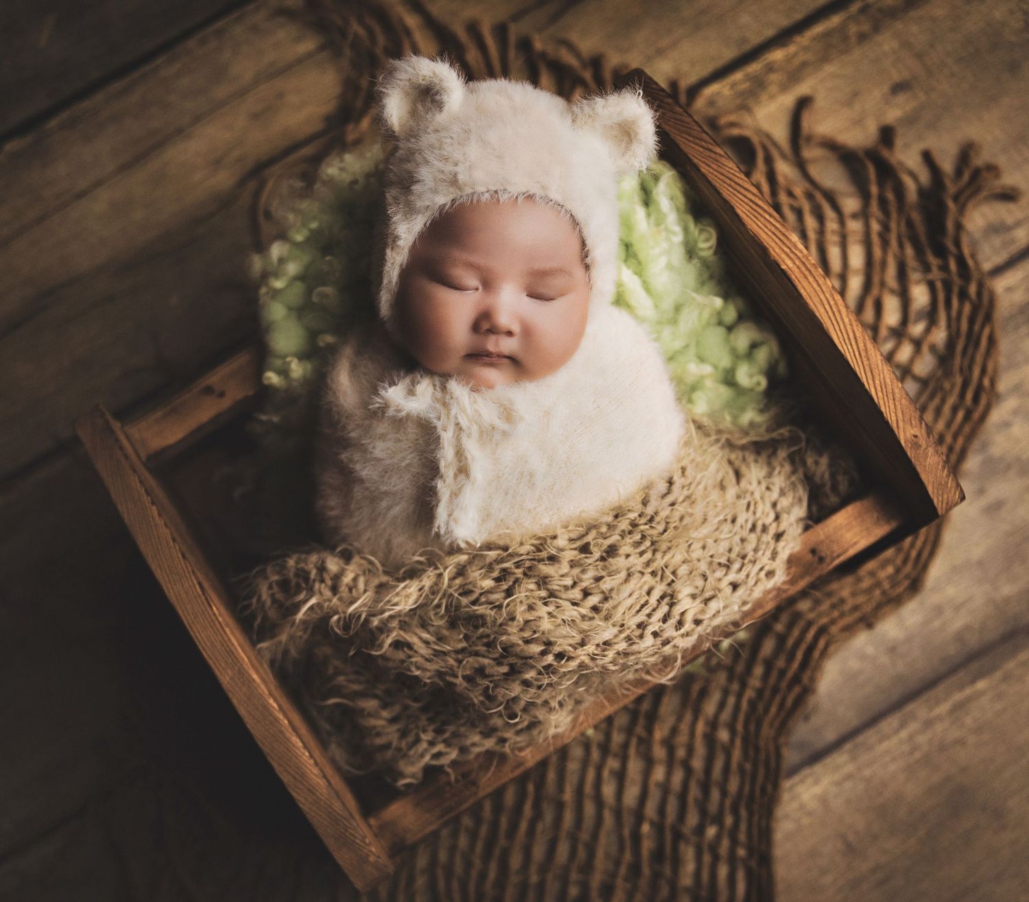 Newborn baby sleeping peacefully in a wooden crate, wearing a white bear bonnet, wrapped in cozy knit blankets.