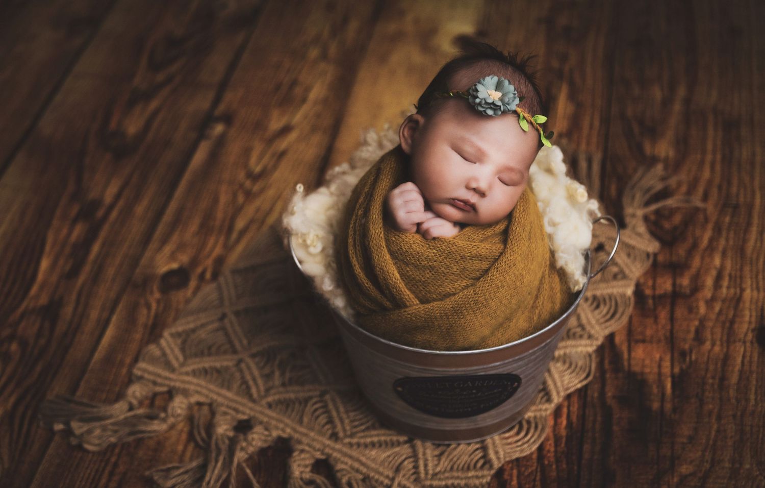 Newborn baby swaddled in mustard yellow wrap with floral headband sleeping in a rustic metal bucket on wood floor.