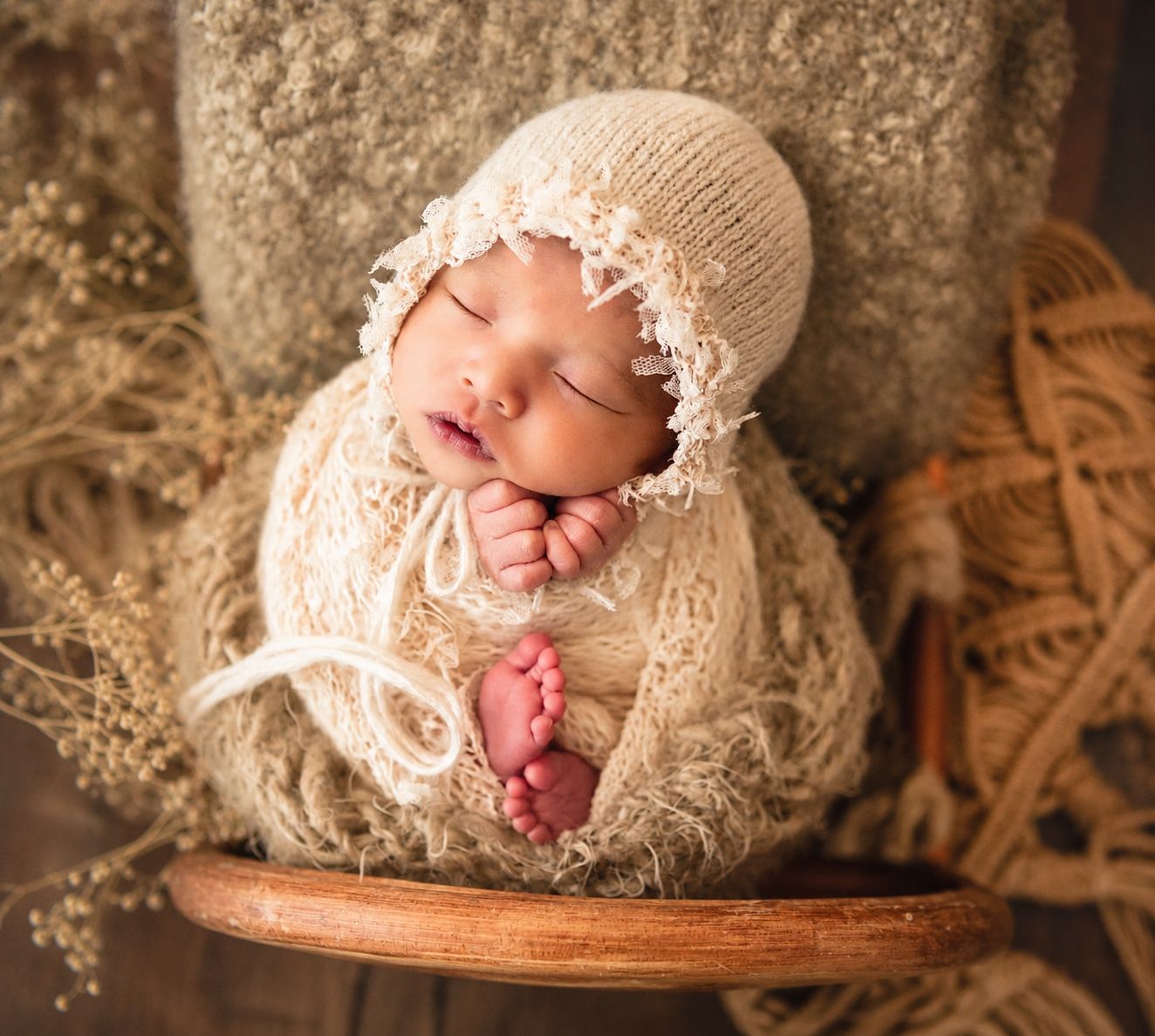 Sleeping newborn baby in cream knit wrap and bonnet, posed in wooden bowl with dried flowers.