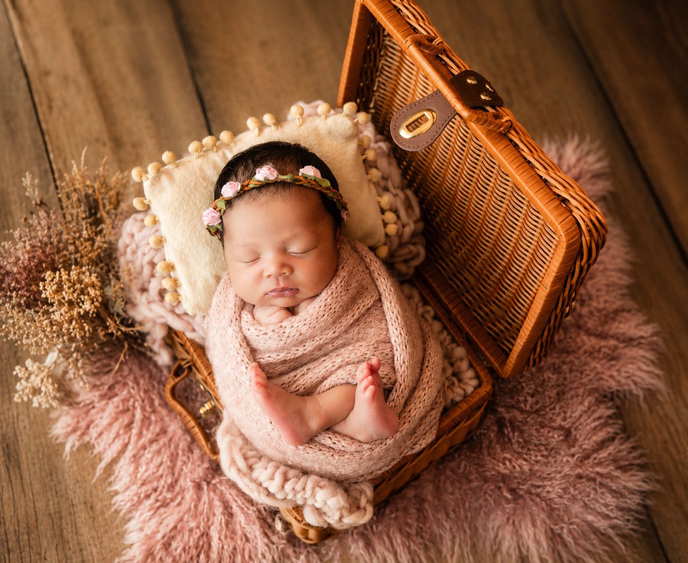 Newborn baby sleeping peacefully wrapped in pink, nestled in a wicker suitcase with a floral headband.