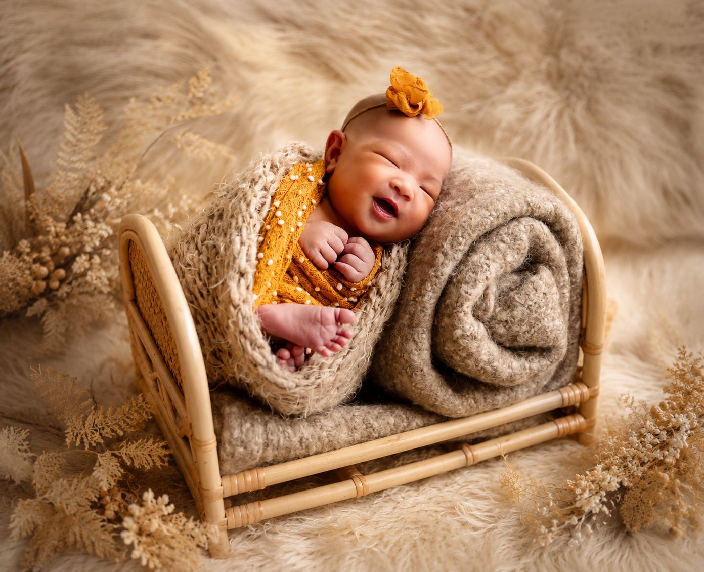 Smiling newborn baby in yellow outfit resting on a tiny wooden bed with fluffy blanket and floral backdrop.
