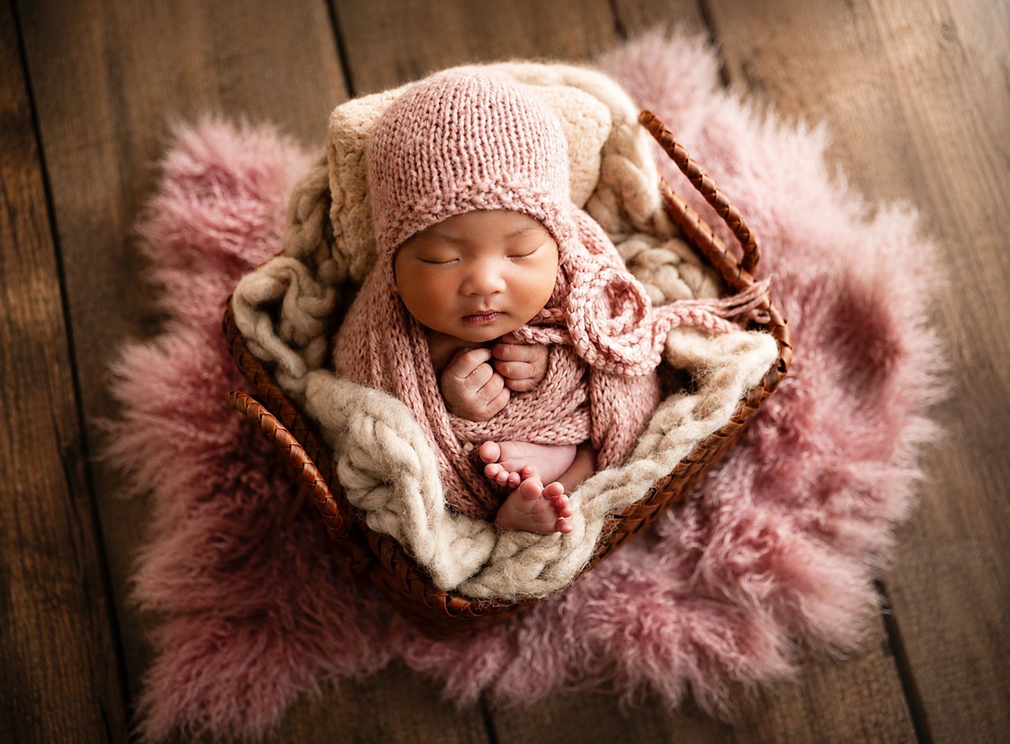 Sleeping newborn baby in pink knit outfit and bonnet nestled in a fluffy pink fur basket on wood floor.