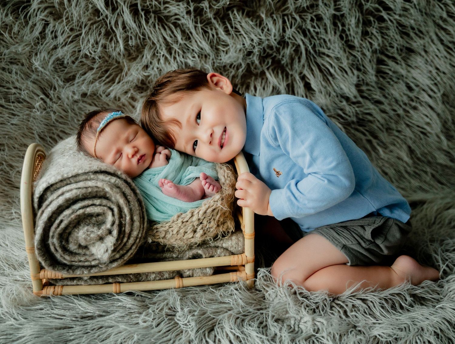 Toddler boy in blue shirt smiles beside sleeping newborn baby in tiny rattan bed on gray fluffy rug.