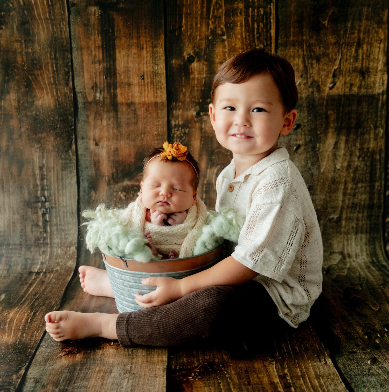 Young boy smiling beside a newborn baby in a bucket with green fluff, posed on rustic wood backdrop.