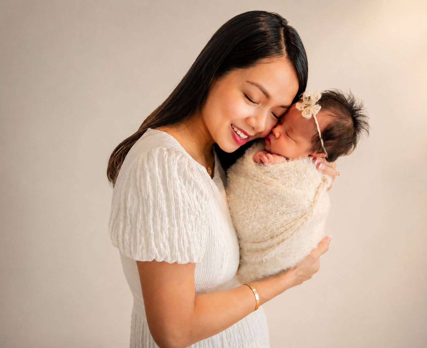 A smiling mother in a white dress tenderly holds her swaddled newborn baby close to her cheek.