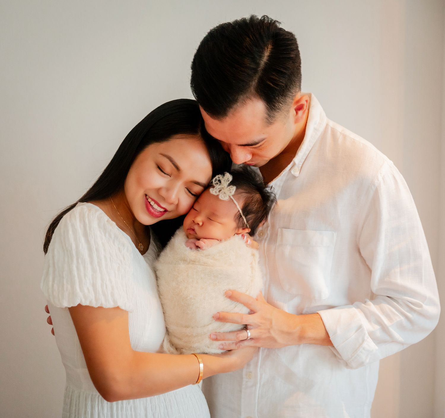 A smiling couple in white outfits tenderly holds their swaddled newborn baby with a tiny bow headband.
