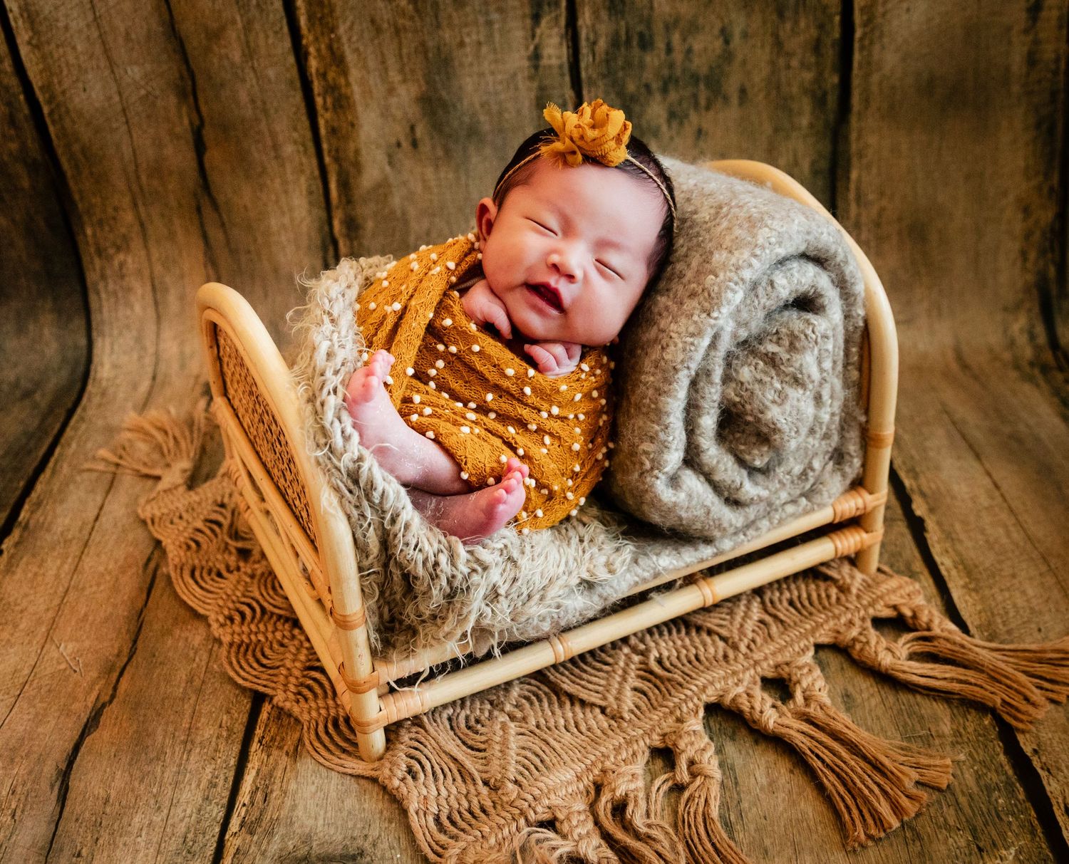 Smiling newborn baby in mustard outfit sleeping on tiny macrame bed with cozy blanket, rustic wood background.