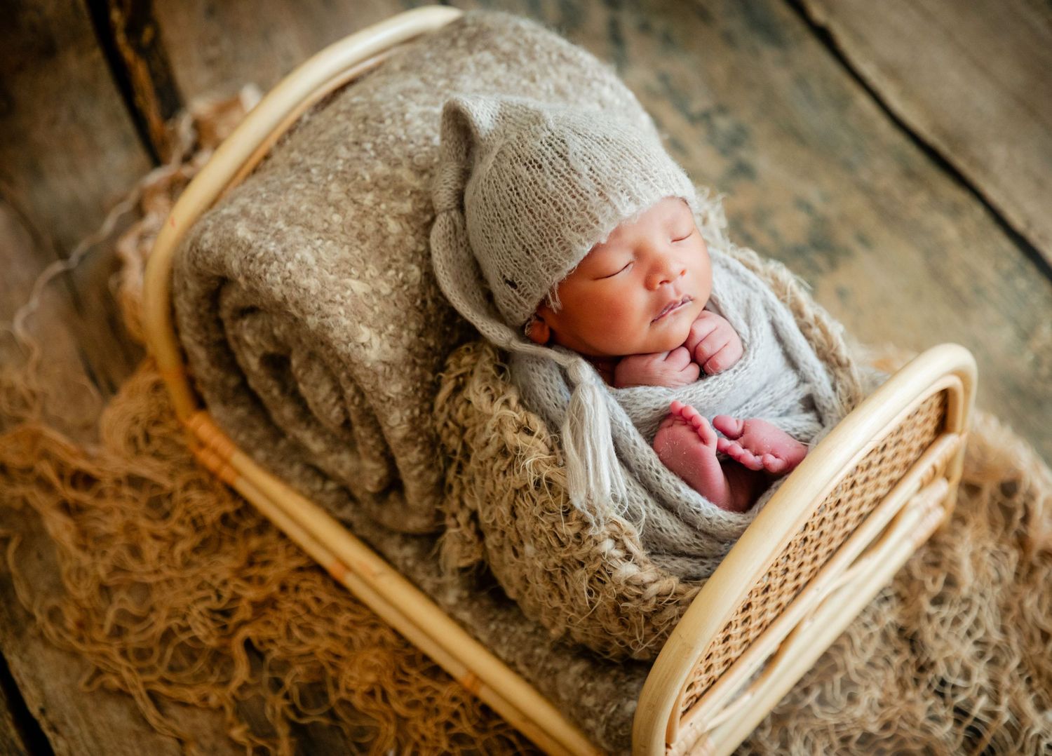 Newborn baby sleeping peacefully in a wicker basket bed, wrapped in gray knit blanket and bear ear hat.