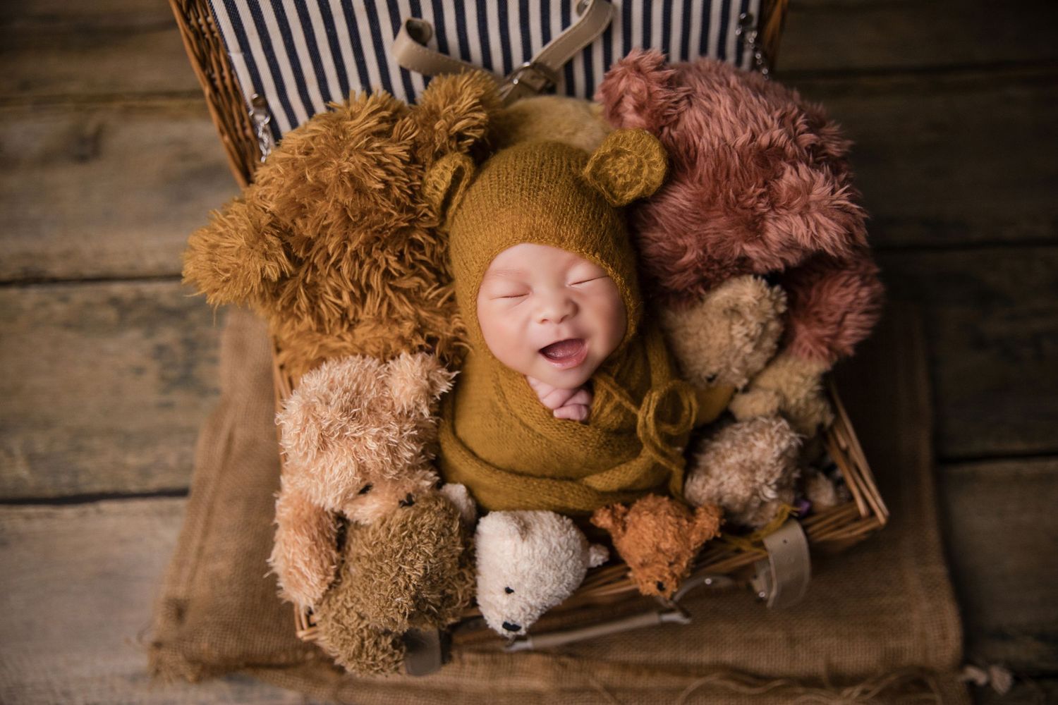 Smiling newborn baby wrapped in mustard yellow, nestled in a basket surrounded by soft teddy bears.