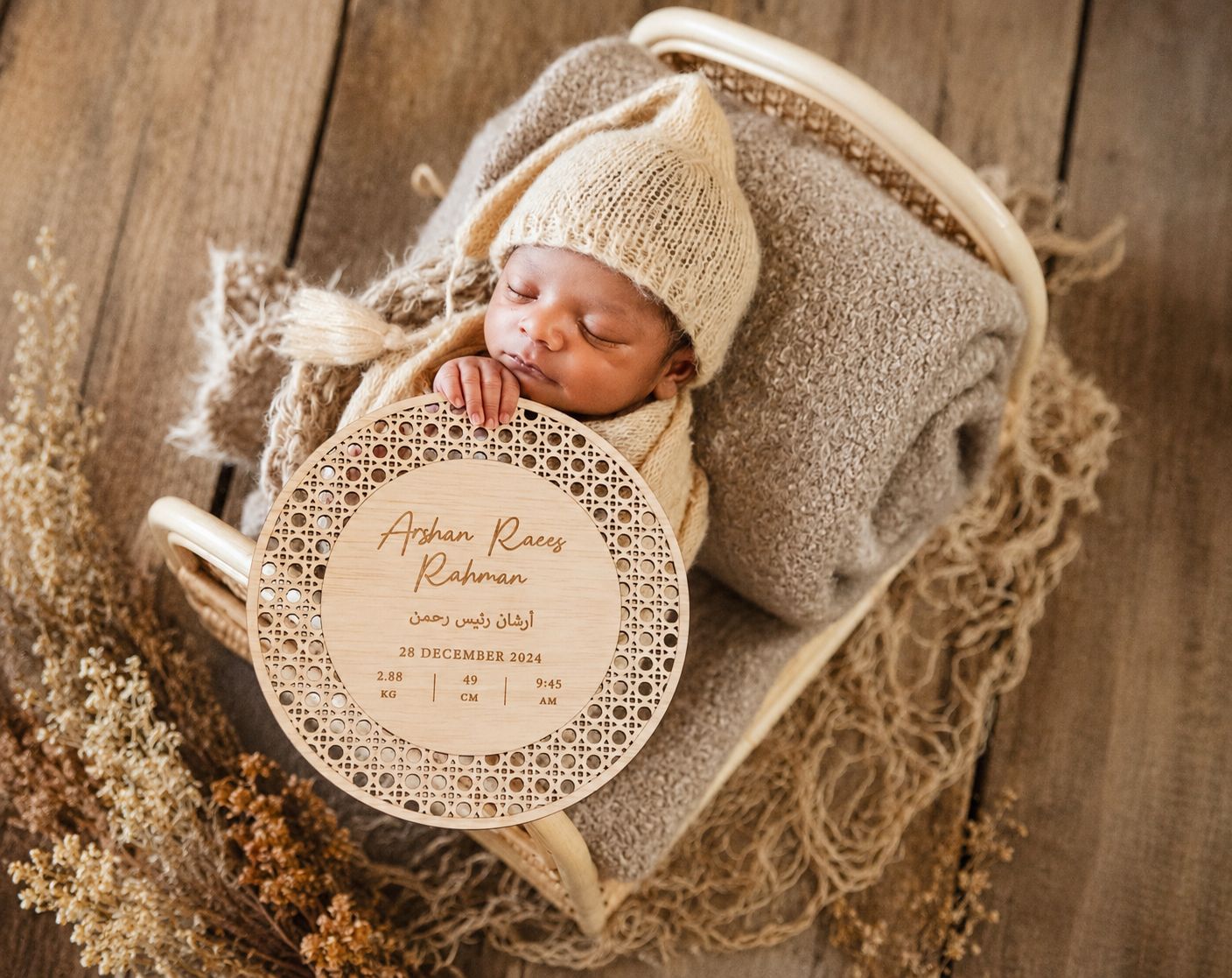 Newborn baby sleeping in a wicker basket with a personalized wooden birth announcement plaque and knit hat.