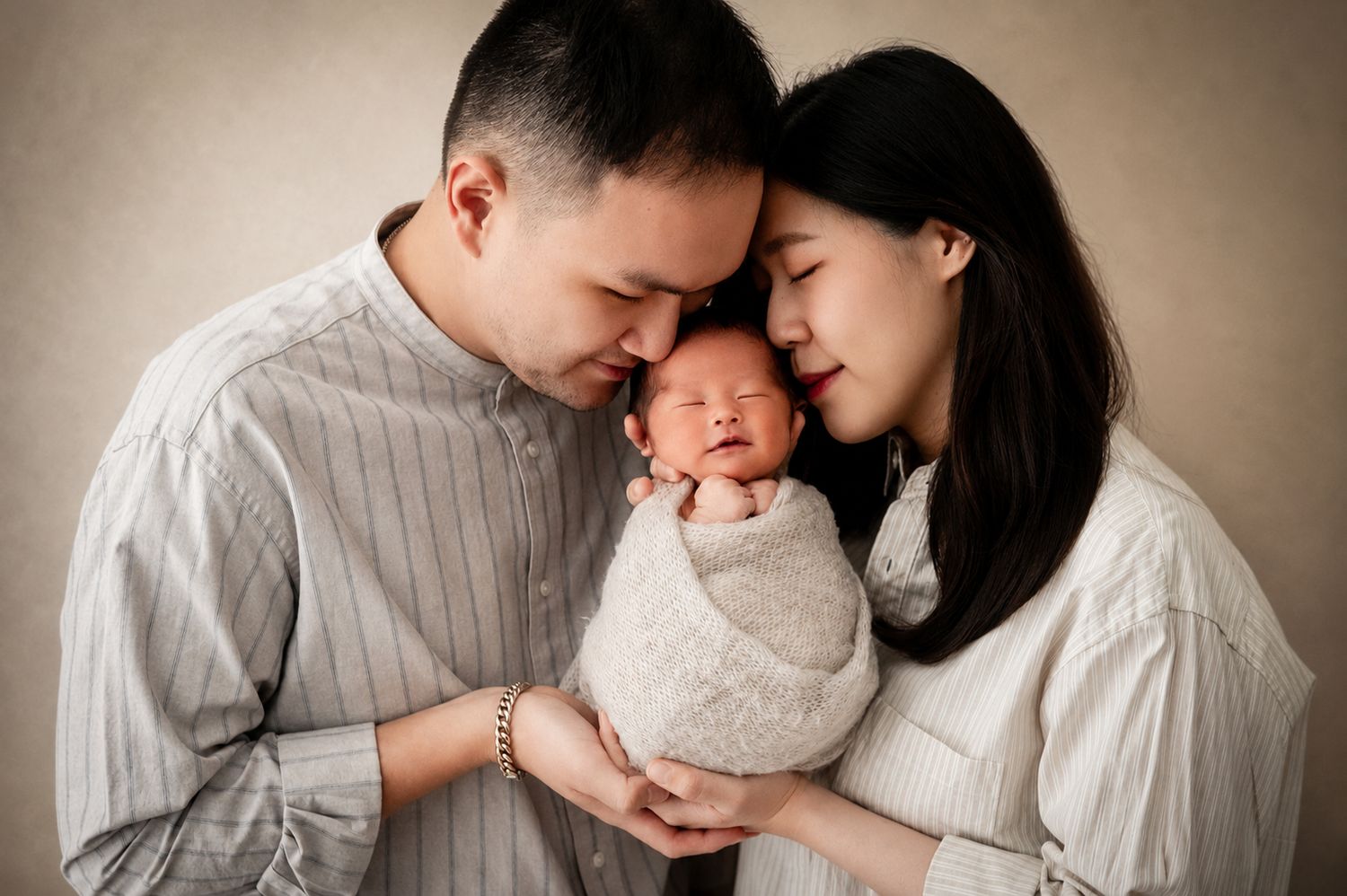Asian couple tenderly holding their swaddled newborn baby together in a warm studio portrait photo.