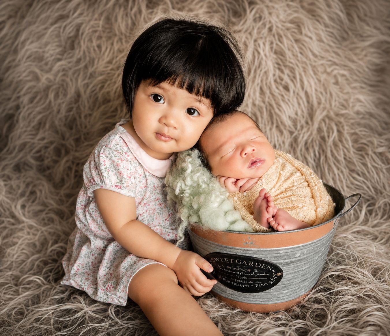 Toddler girl in floral dress poses with sleeping newborn baby wrapped in lace inside a vintage metal bucket.