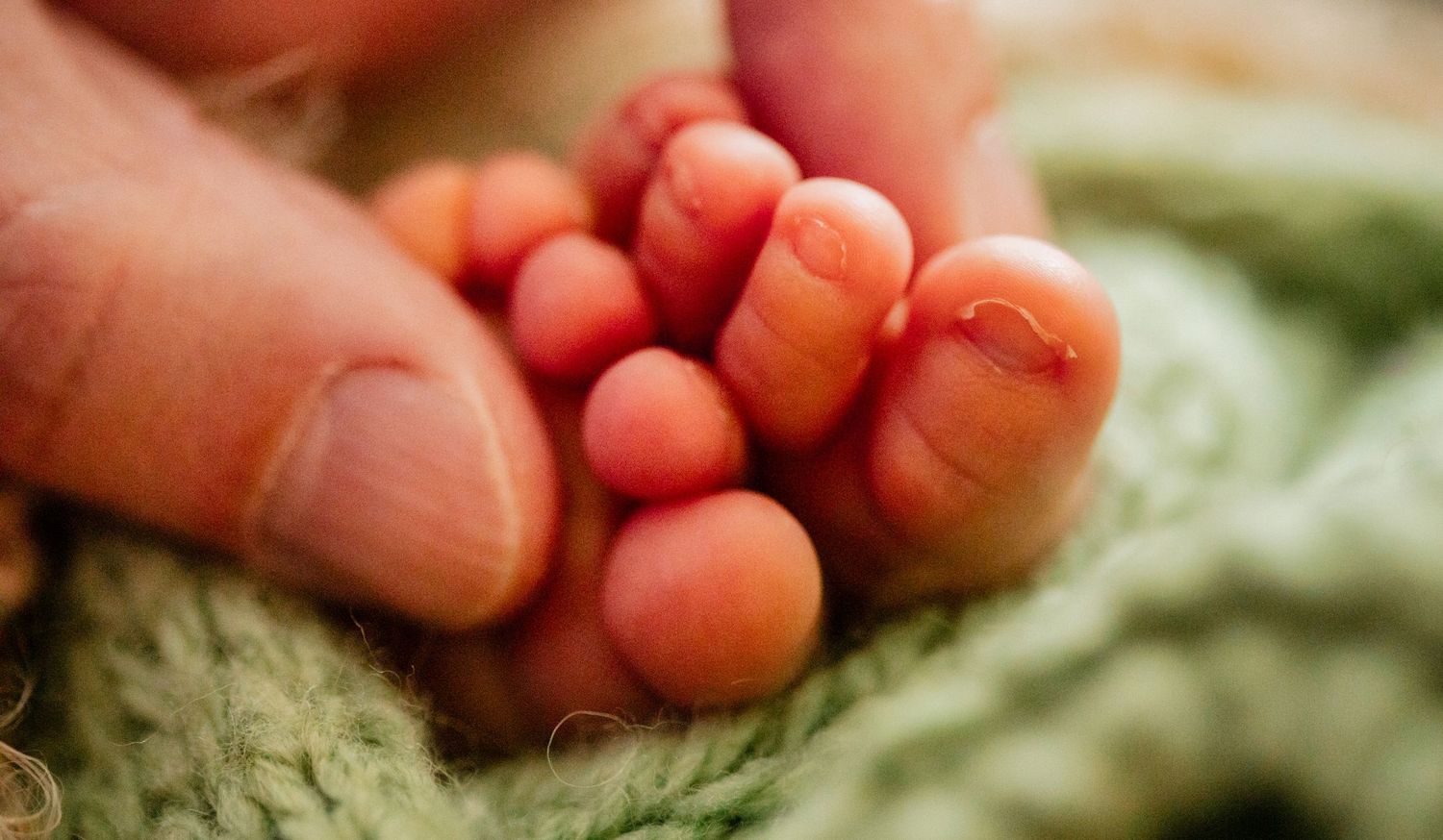 Close-up of a newborn baby's tiny toes held gently by an adult hand, wrapped in a soft green knit blanket.