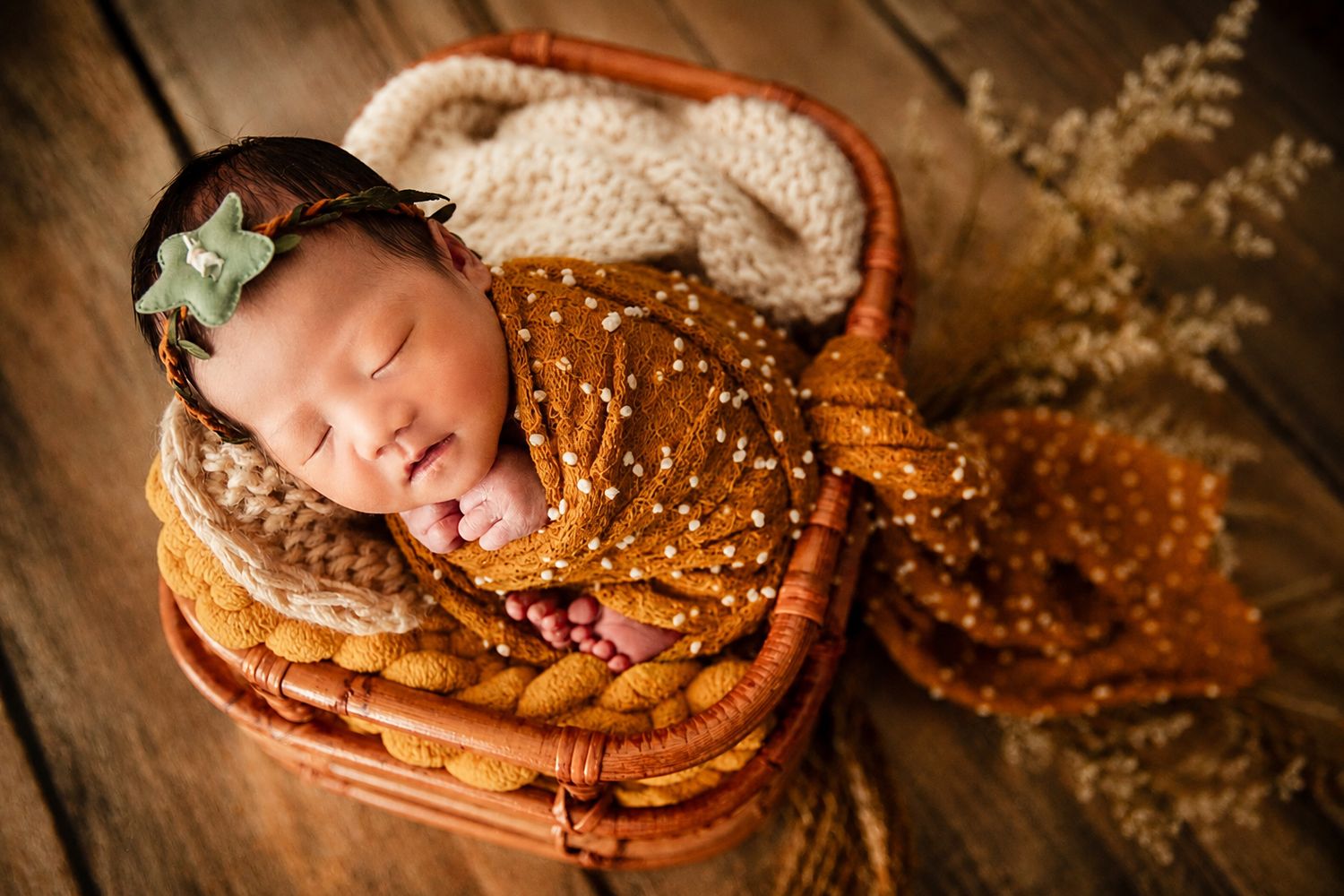 Sleeping newborn baby wrapped in rust orange swaddle with green bow headband nestled in wicker basket.