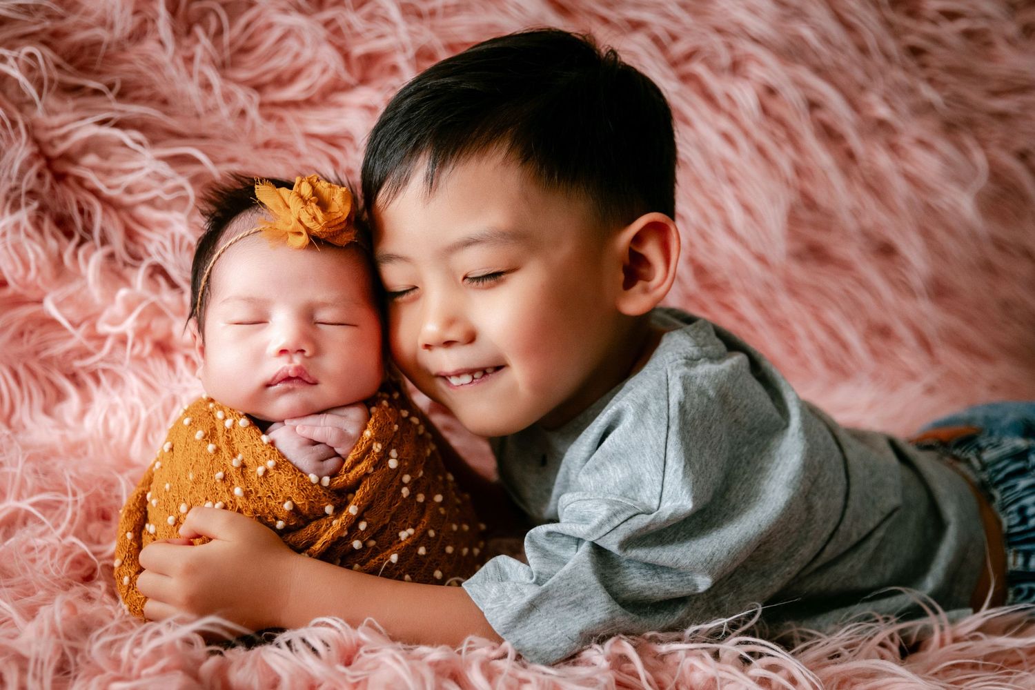 A smiling young boy gently hugs a sleeping newborn baby girl with a bow on a pink fluffy blanket.