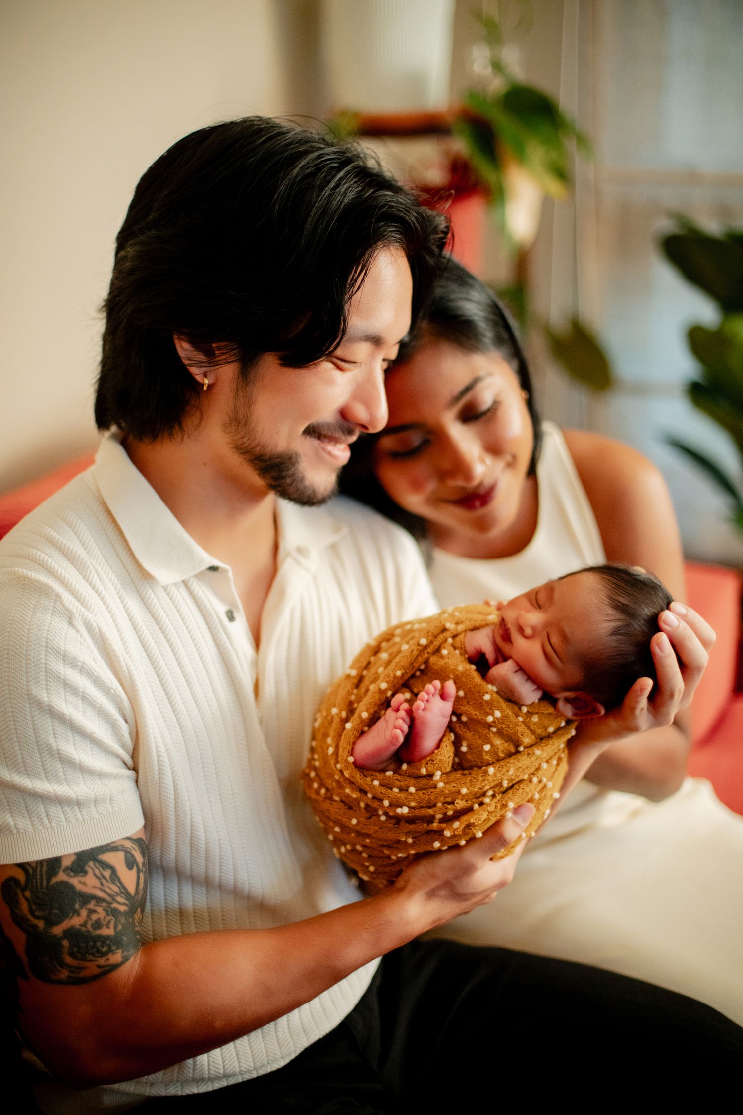 Young couple smiling and holding their sleeping newborn baby wrapped in a golden blanket at home.