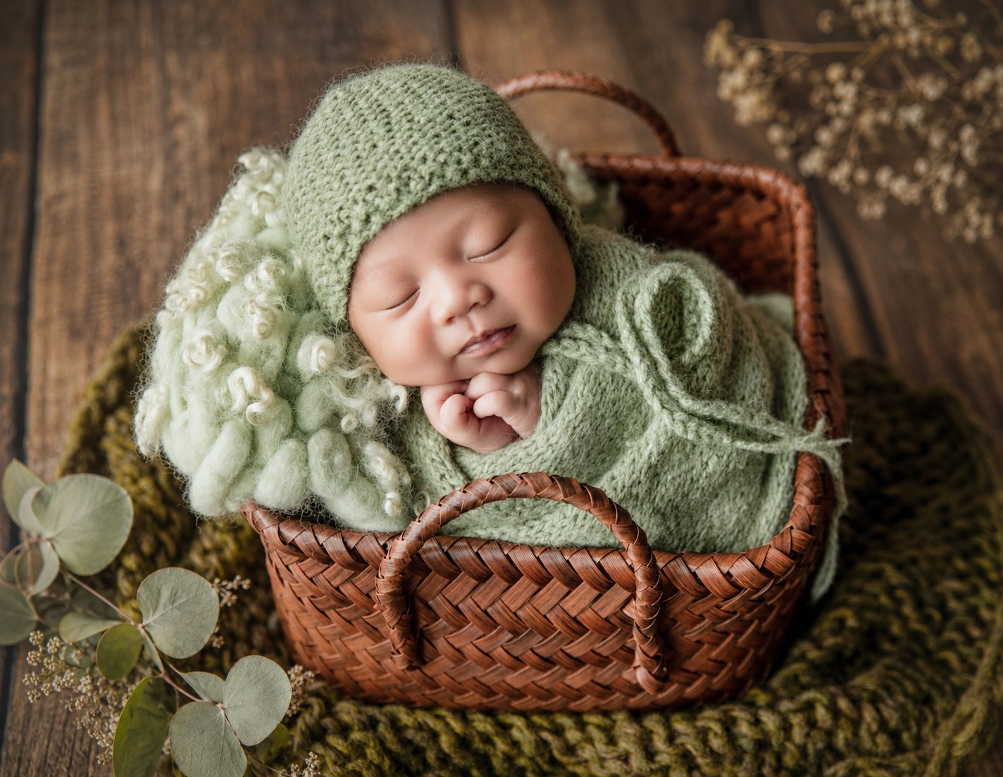 Newborn baby sleeping in wicker basket wrapped in green knit blanket and bonnet surrounded by white flowers.