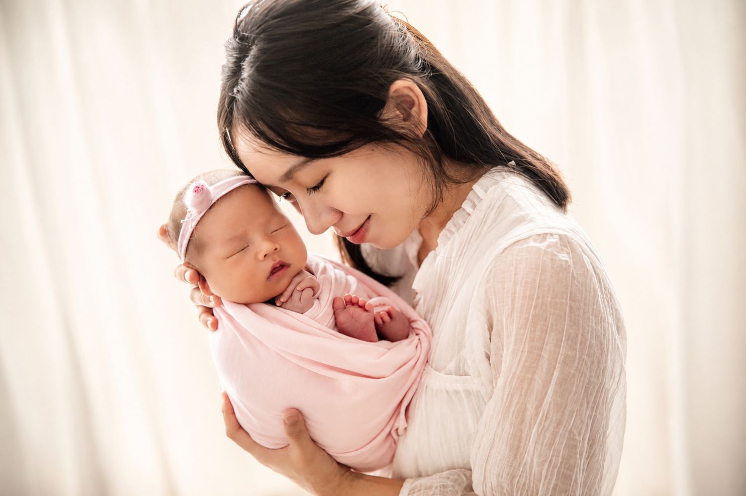 A young Asian mother tenderly holds her sleeping newborn baby wrapped in a pink swaddle with a bow headband.