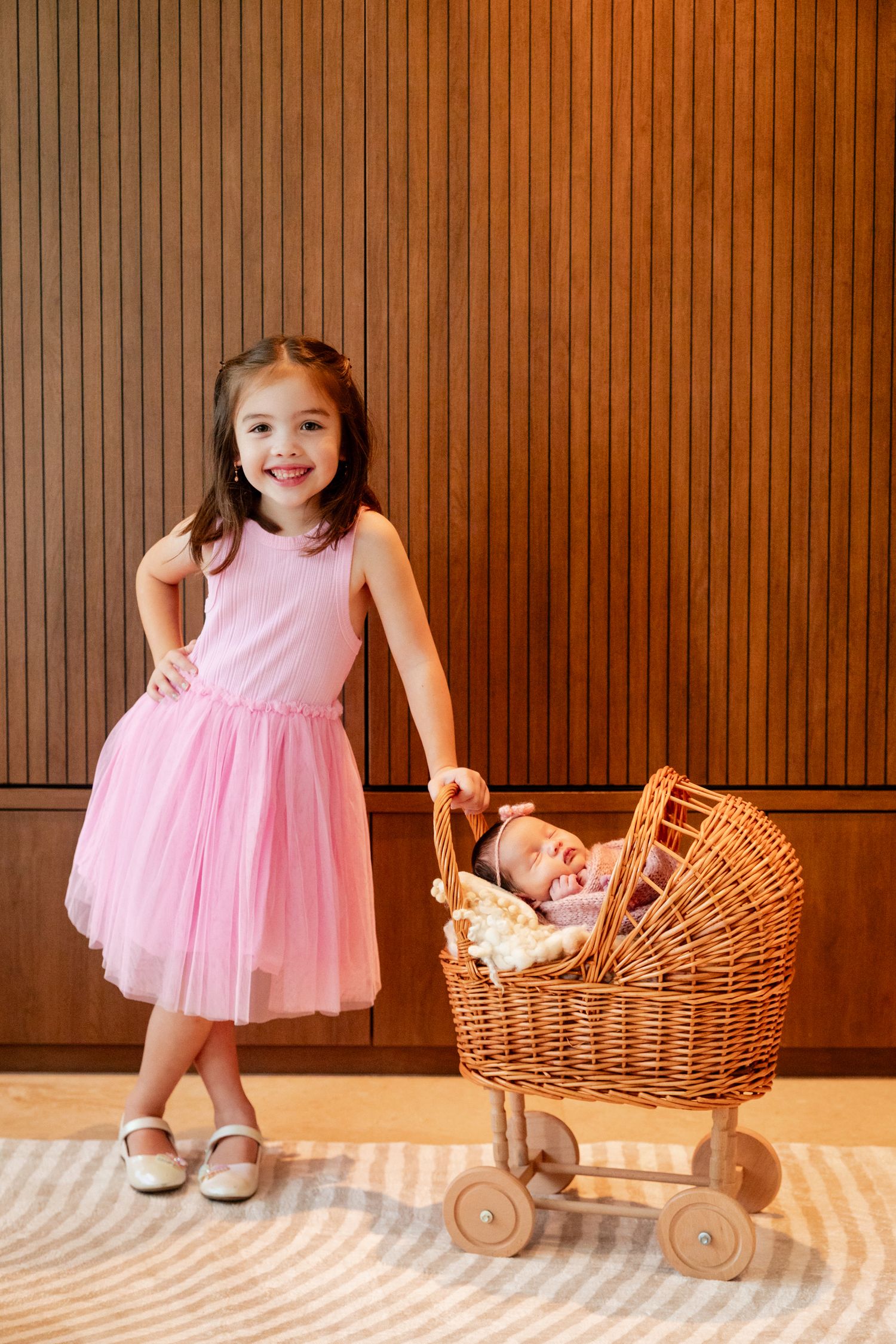 A smiling girl in a pink tutu dress stands beside a wicker baby carriage holding a sleeping newborn indoors.