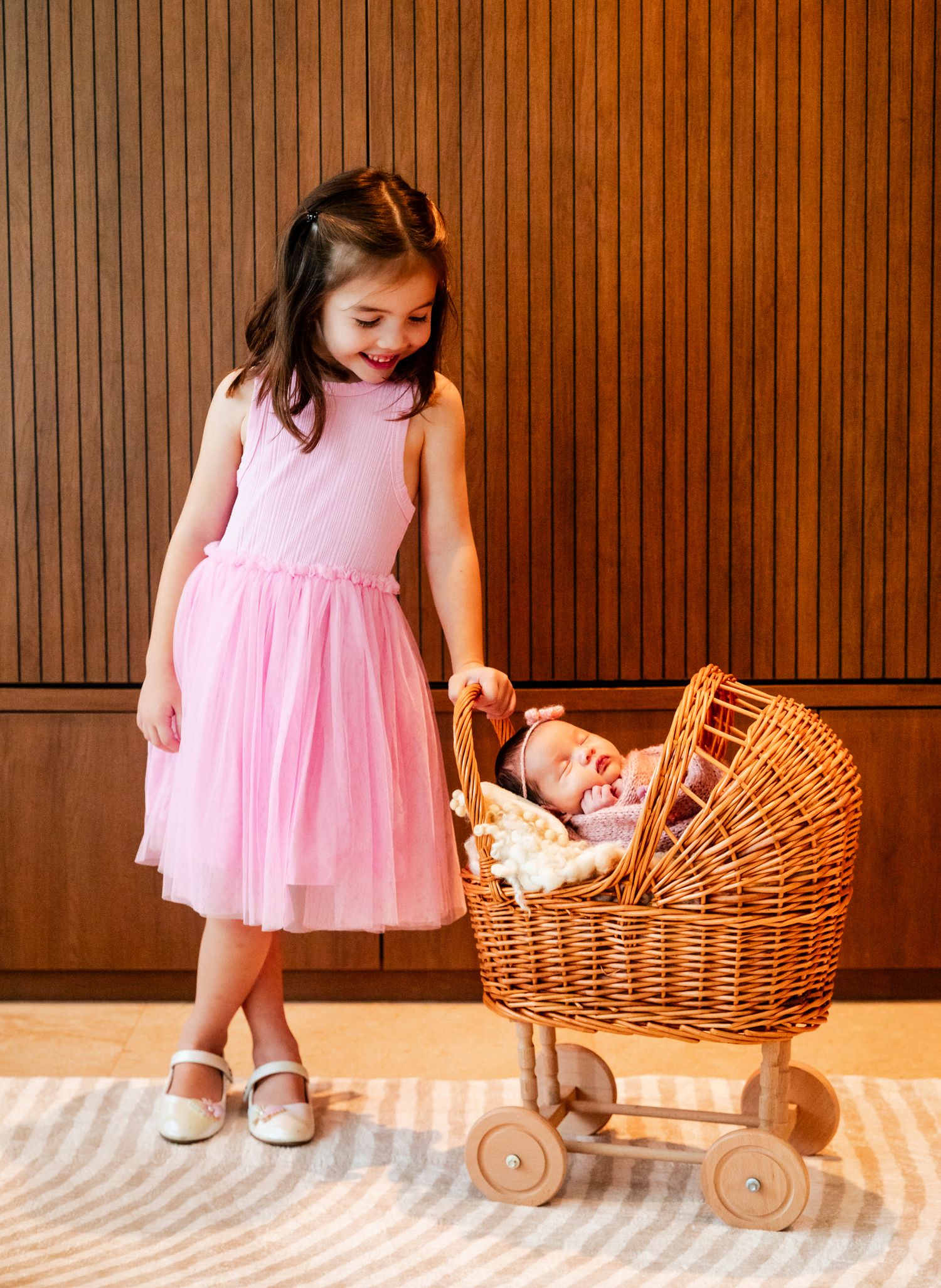 A young girl in a pink tulle dress smiles at a newborn baby resting in a wicker doll pram on a rug.