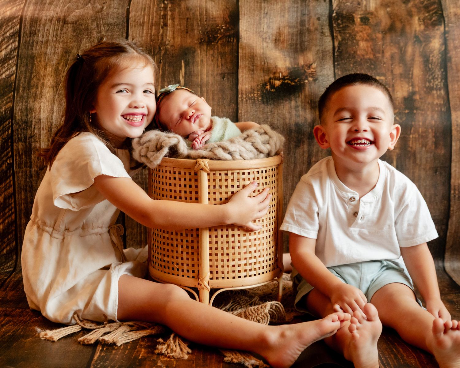 Two smiling children pose with a sleeping newborn baby in a wicker basket against a rustic wood background.