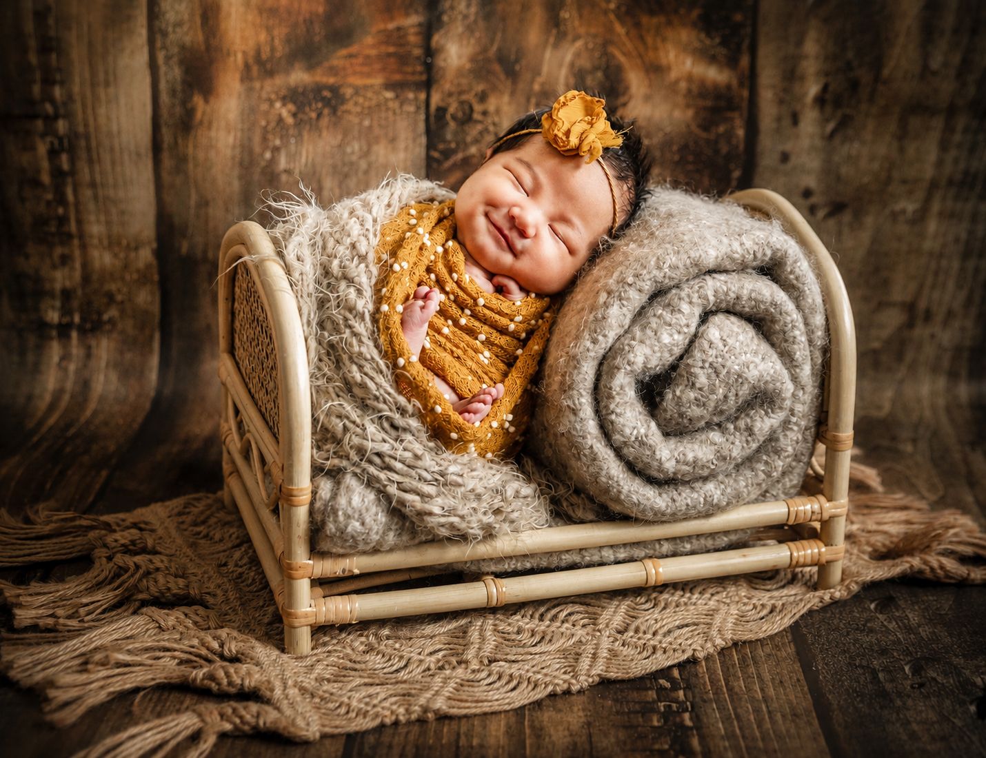 Smiling newborn baby sleeping in a small rustic wooden bed with cozy gray blanket and yellow floral headband.