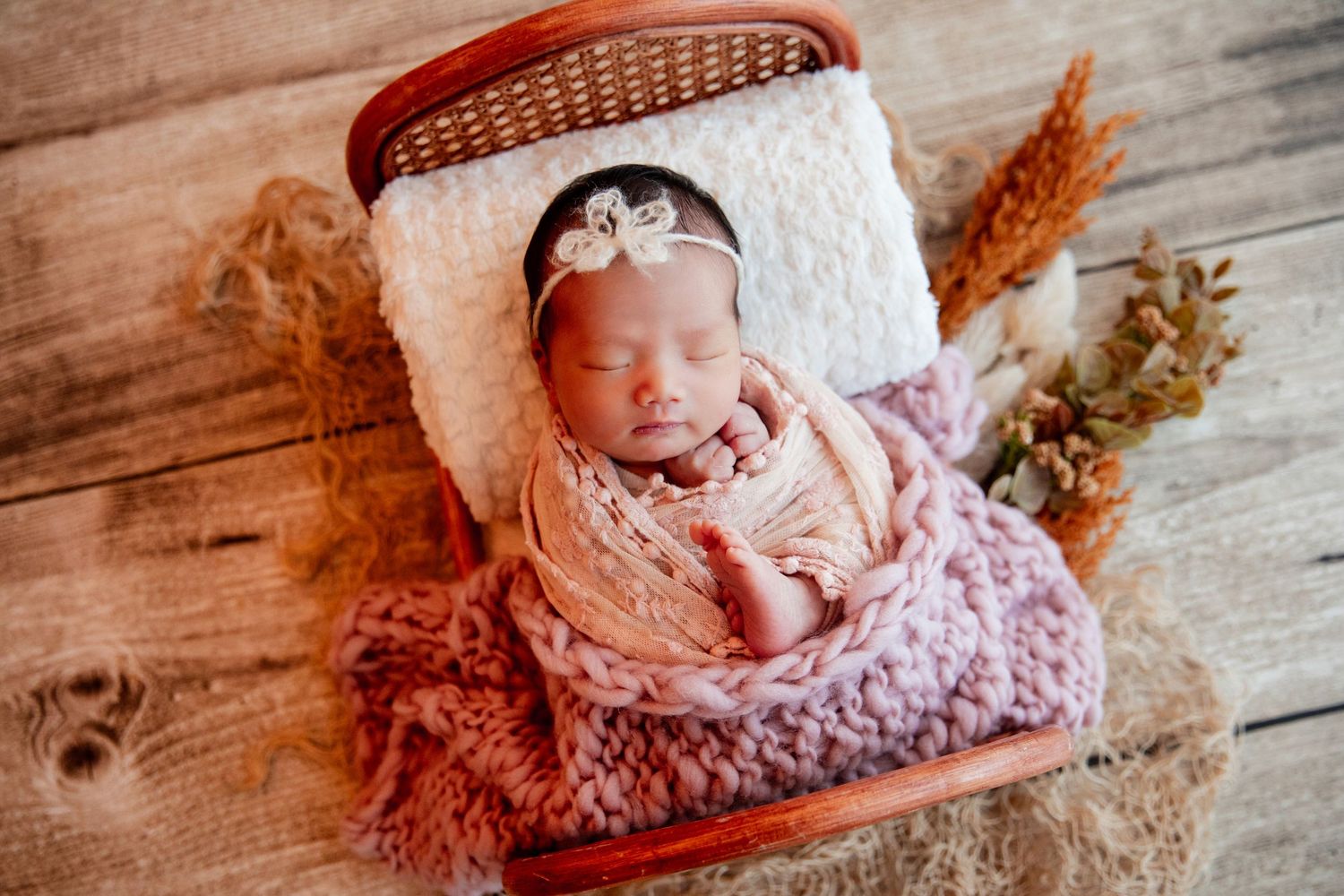 Newborn baby sleeping peacefully in a wicker basket, wrapped in pink and mauve knit blankets with a floral headband.