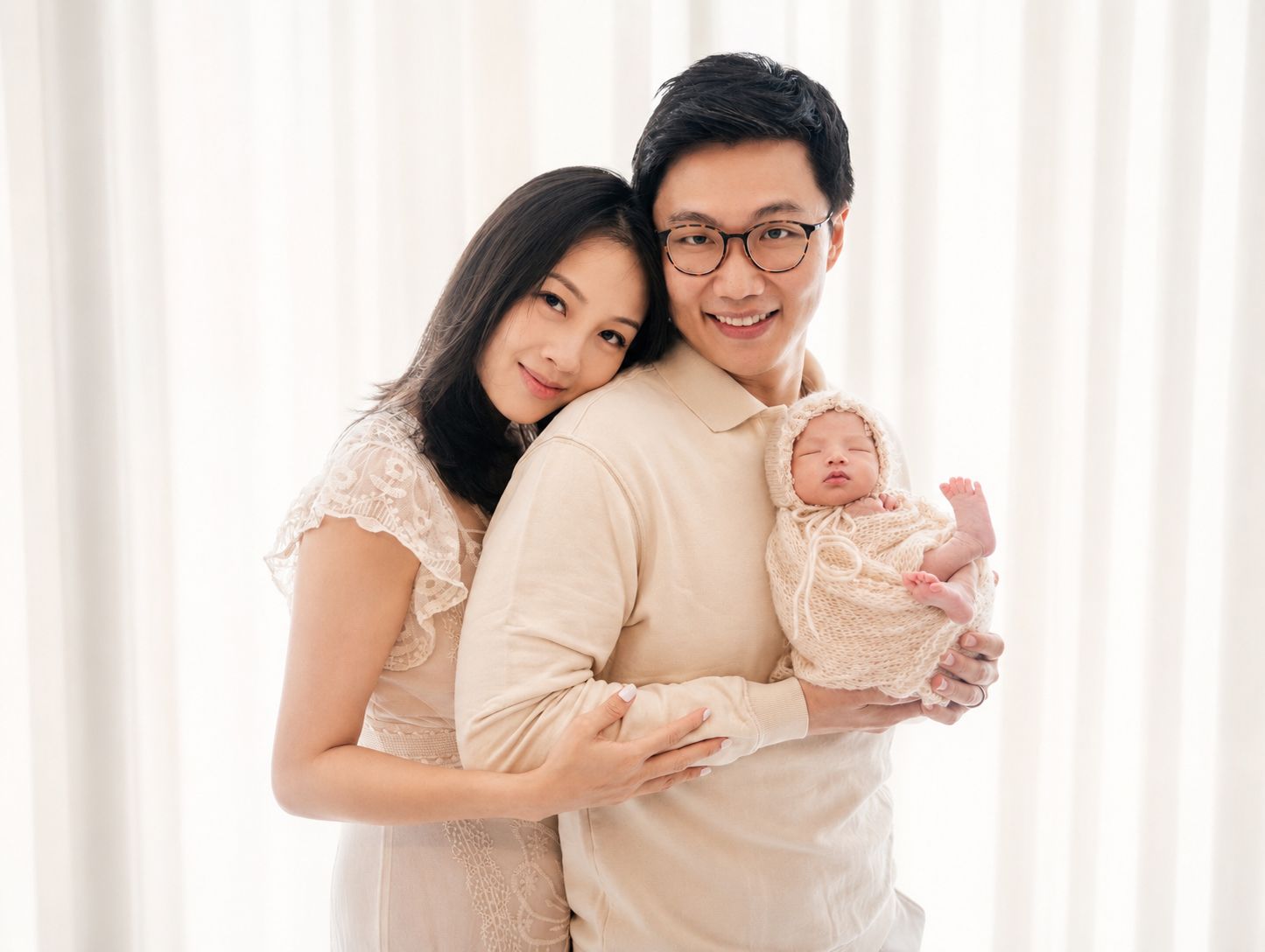 A happy Asian couple in neutral tones poses with their newborn baby against a soft white curtain backdrop.