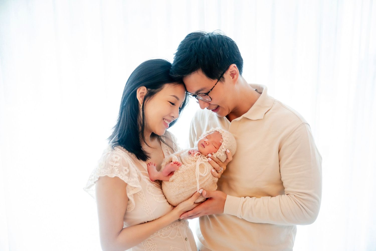 A couple tenderly holds their newborn baby together, smiling lovingly in a soft, bright studio setting.