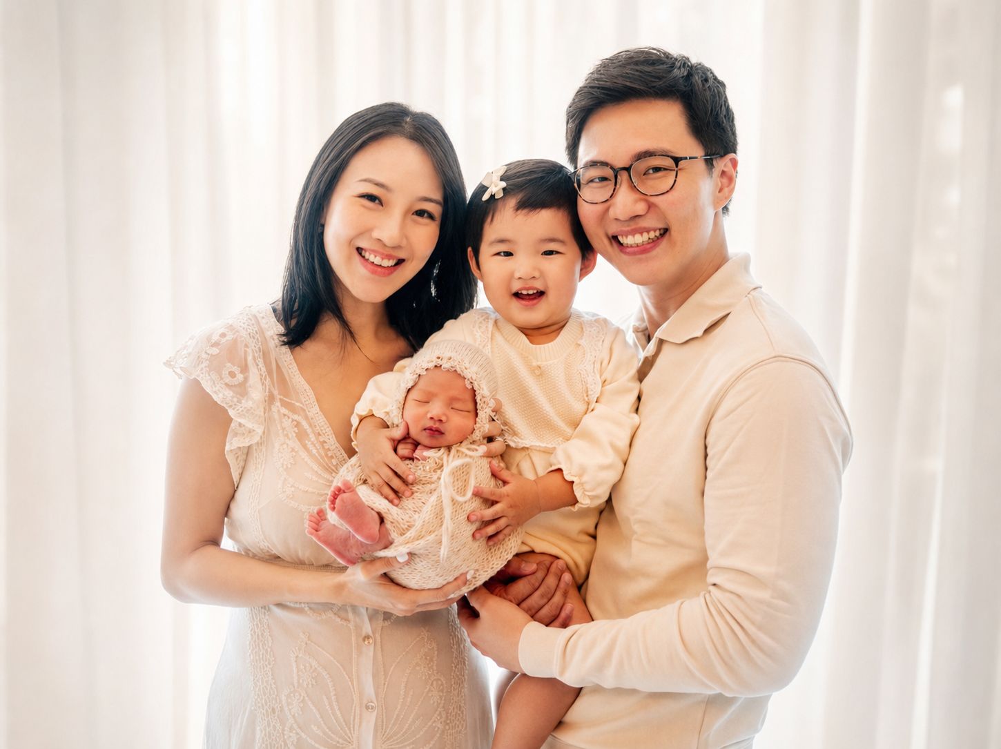 Happy Asian family posing for newborn photo shoot with toddler daughter and baby in white studio setting.