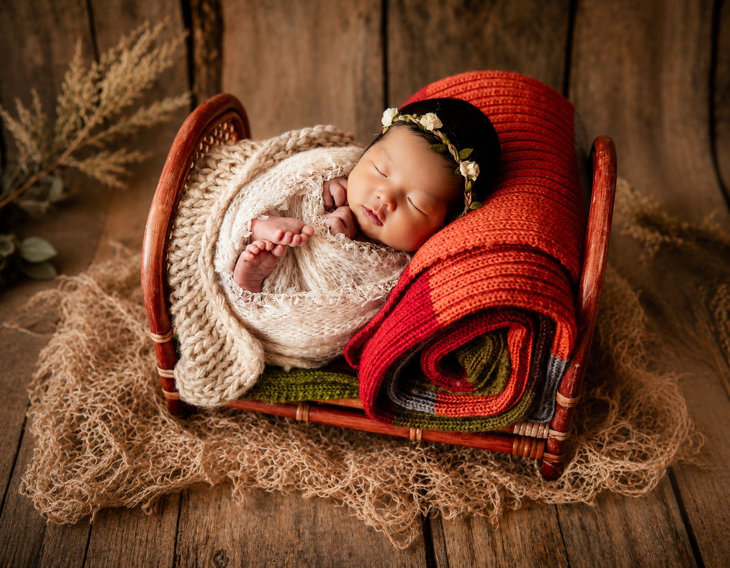 Sleeping newborn baby wrapped in cream knit swaddle resting in a red heart-shaped wicker basket with colorful blankets.