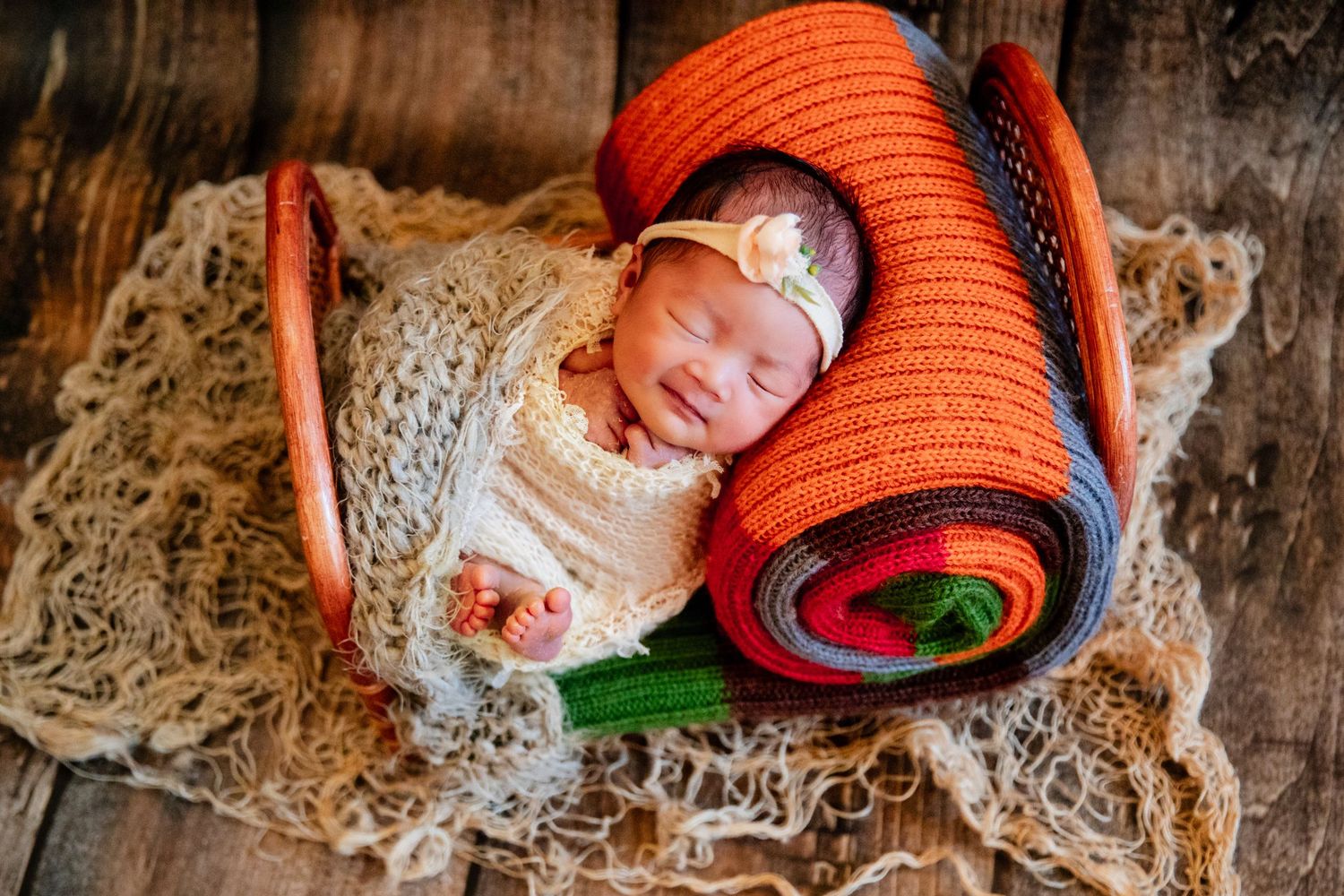 Sleeping newborn baby wrapped in cream swaddle with floral headband nestled in basket with colorful rolled blankets.