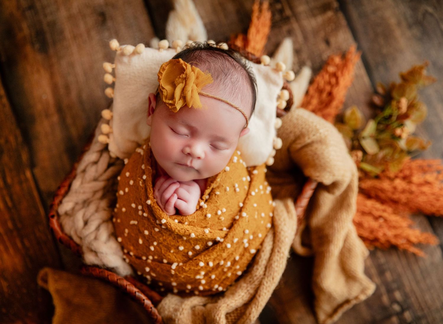 Newborn baby girl swaddled in mustard wrap with floral headband, sleeping peacefully in autumn-themed photo setup.