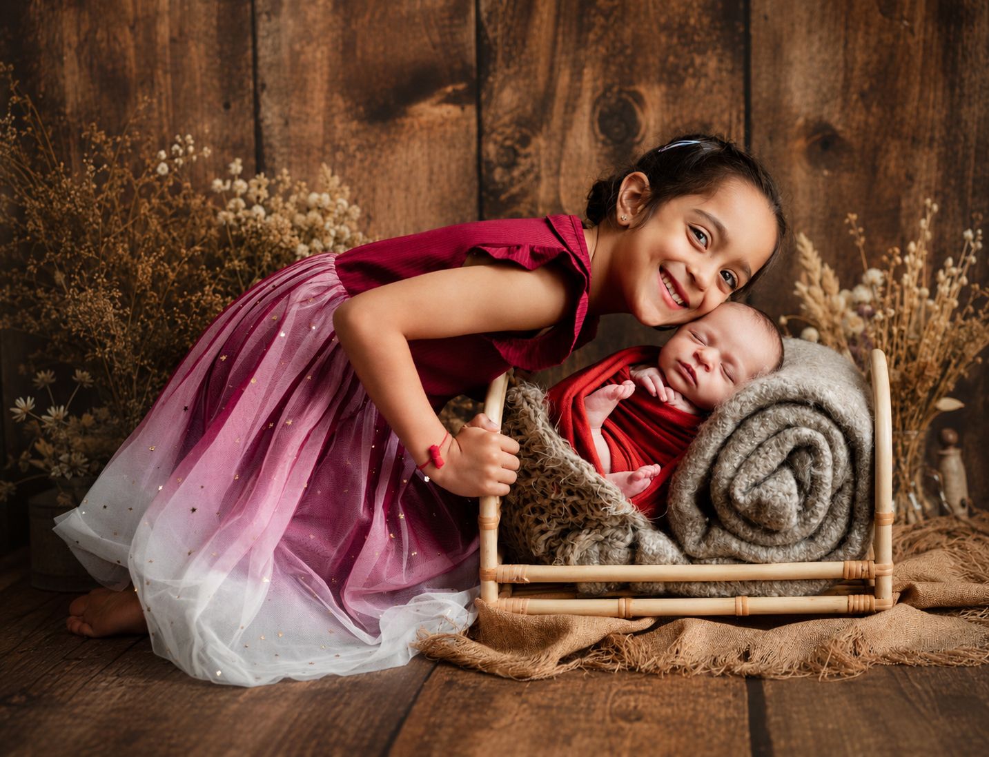 A young girl in a pink dress smiles while hugging a sleeping newborn in a red wrap on a rustic wooden bed.