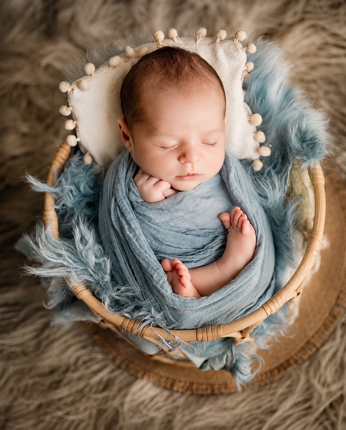 Newborn baby sleeping peacefully in a wicker basket wrapped in a soft blue swaddle with fluffy blue fur lining.