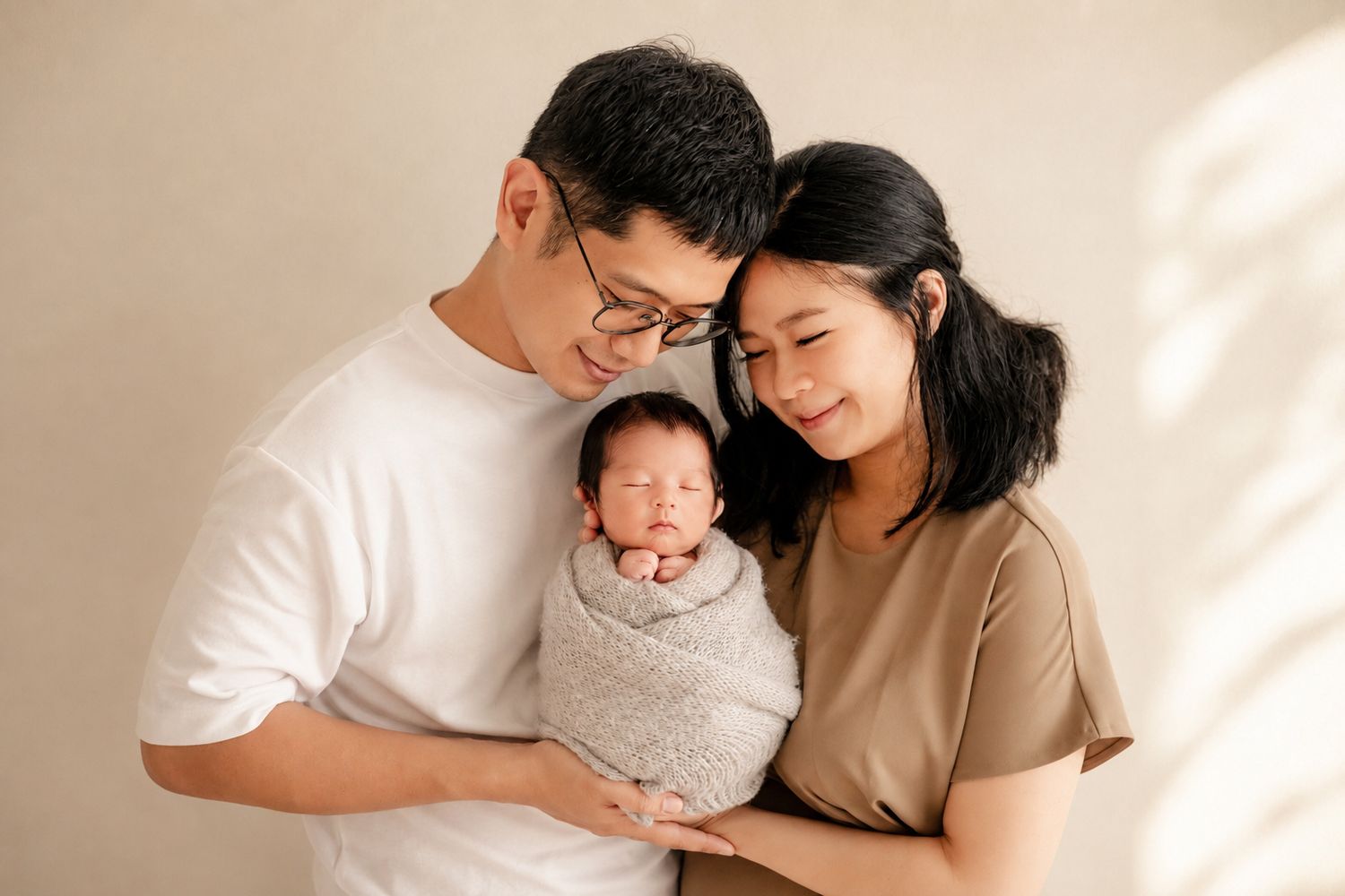 A young Asian couple smiles tenderly while cradling their swaddled newborn baby in a soft, neutral-toned studio setting.