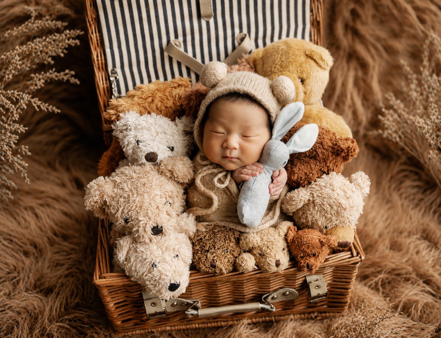 Sleeping newborn baby in knit bonnet nestled in wicker basket surrounded by soft teddy bears and stuffed animals.