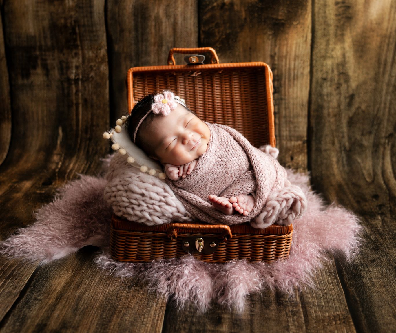 Newborn baby sleeping peacefully in a small wicker suitcase, wrapped in a pink knit blanket with a floral headband.