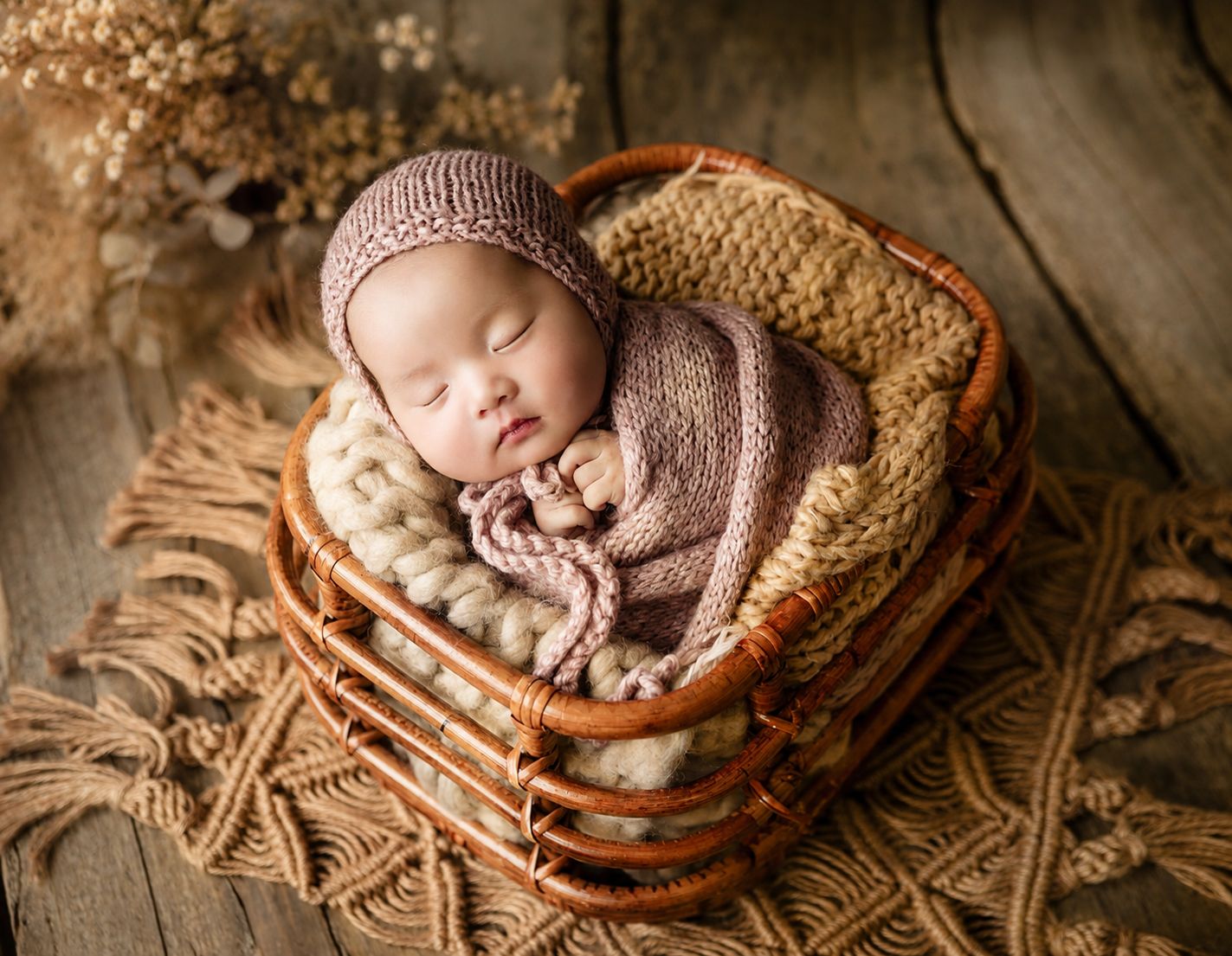 Sleeping newborn baby wrapped in pink knit blanket and bonnet nestled in a wicker basket on rustic wood.