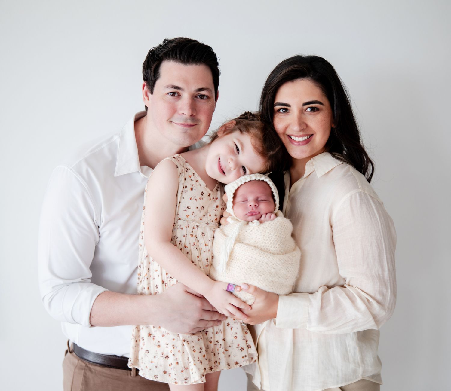 A smiling family of four poses together, parents holding a toddler girl and swaddled newborn baby against a white background.