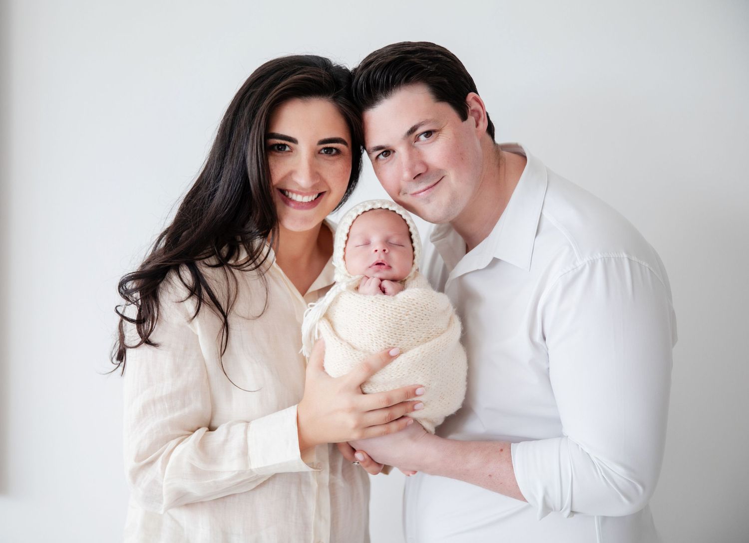Smiling couple in white outfits holding their swaddled newborn baby against a white background.