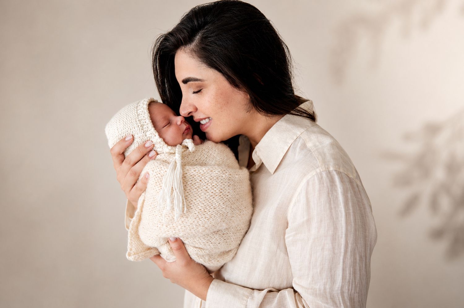 A mother tenderly holds her swaddled newborn baby, both dressed in cream tones against a soft neutral background.