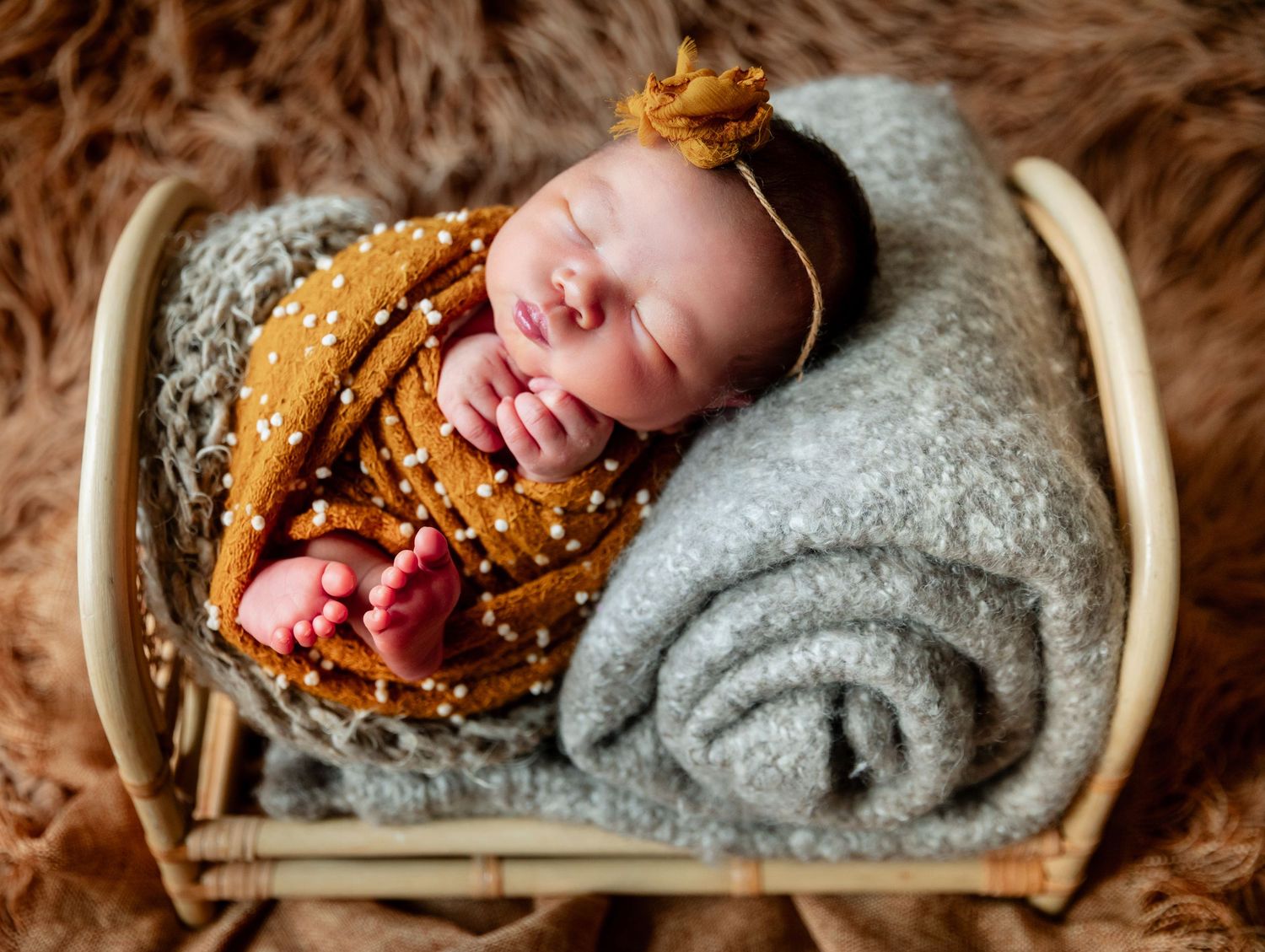 Newborn baby sleeping peacefully in a tiny wicker bed, wrapped in a mustard swaddle with a gold bow headband.