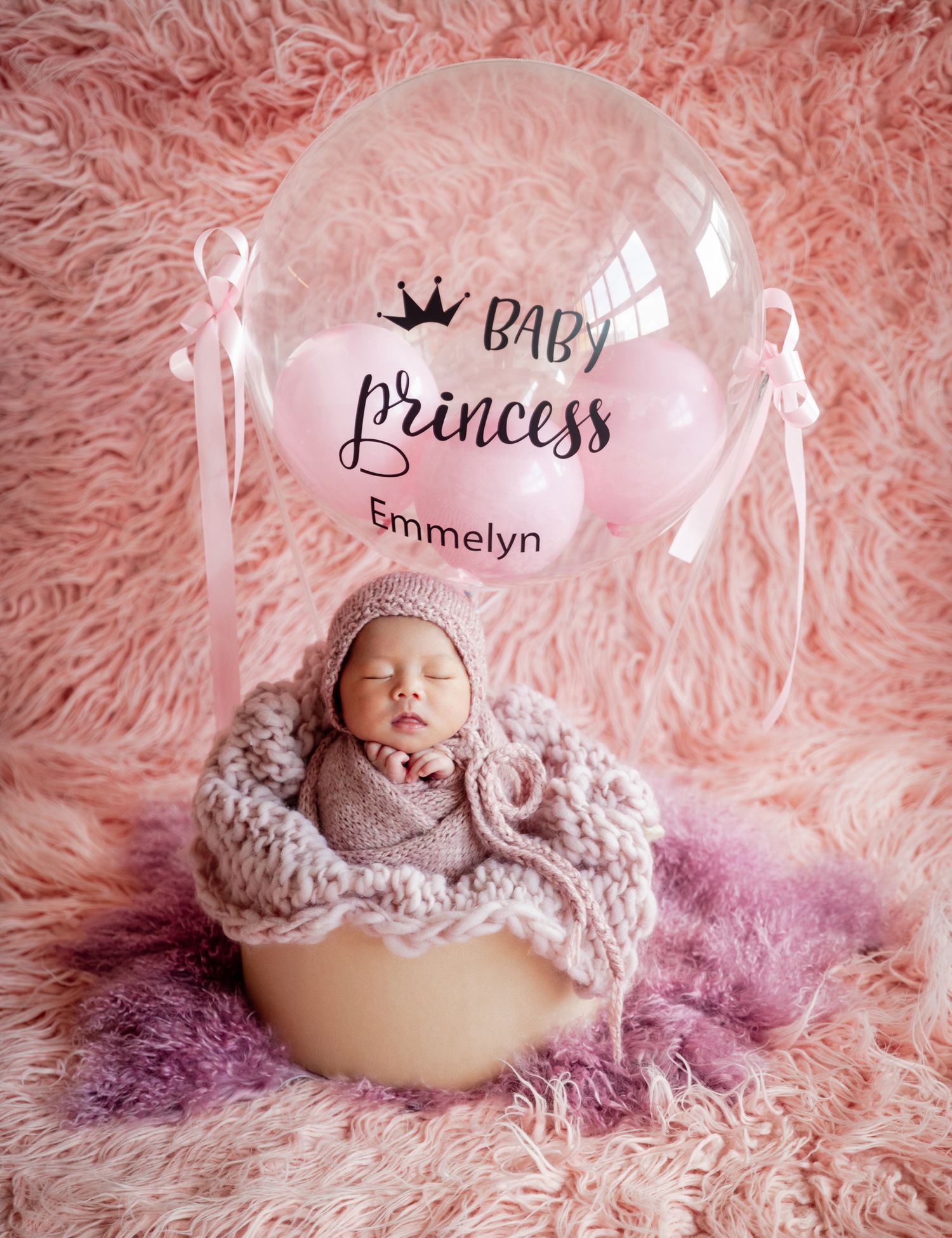 Newborn baby girl named Emmelyn sleeping in a bowl with a pink Baby Princess balloon on a fluffy pink background.