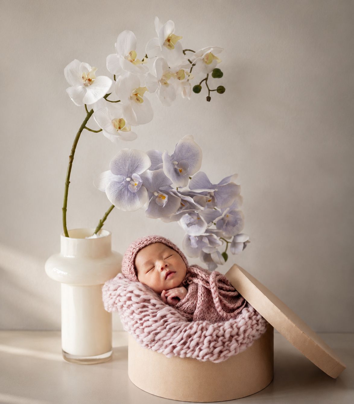 Sleeping newborn baby in pink knit wrap inside a wooden box beside white and purple orchids.