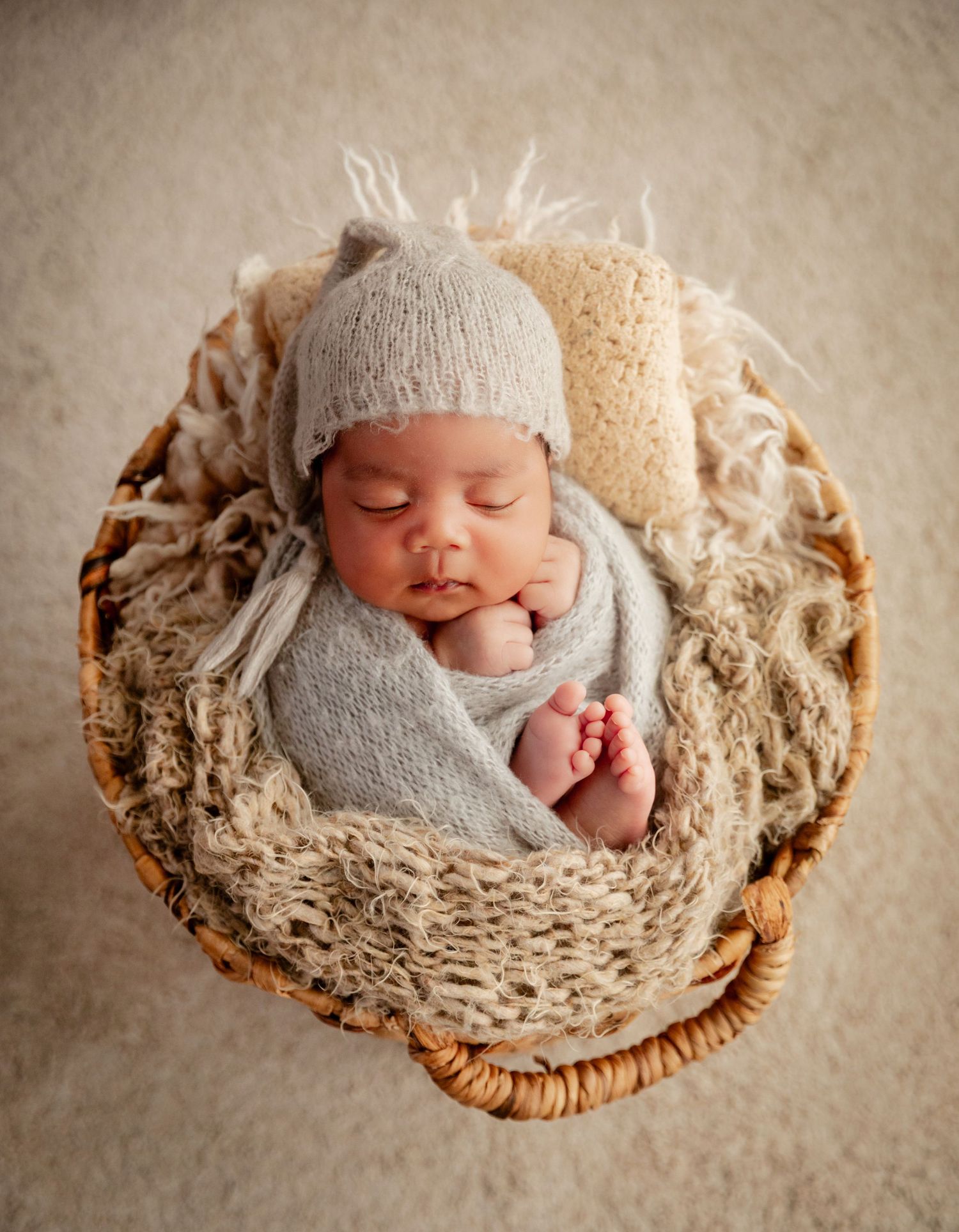 Sleeping newborn baby wrapped in gray swaddle wearing knit hat, nestled in wicker basket with cozy textured blankets.