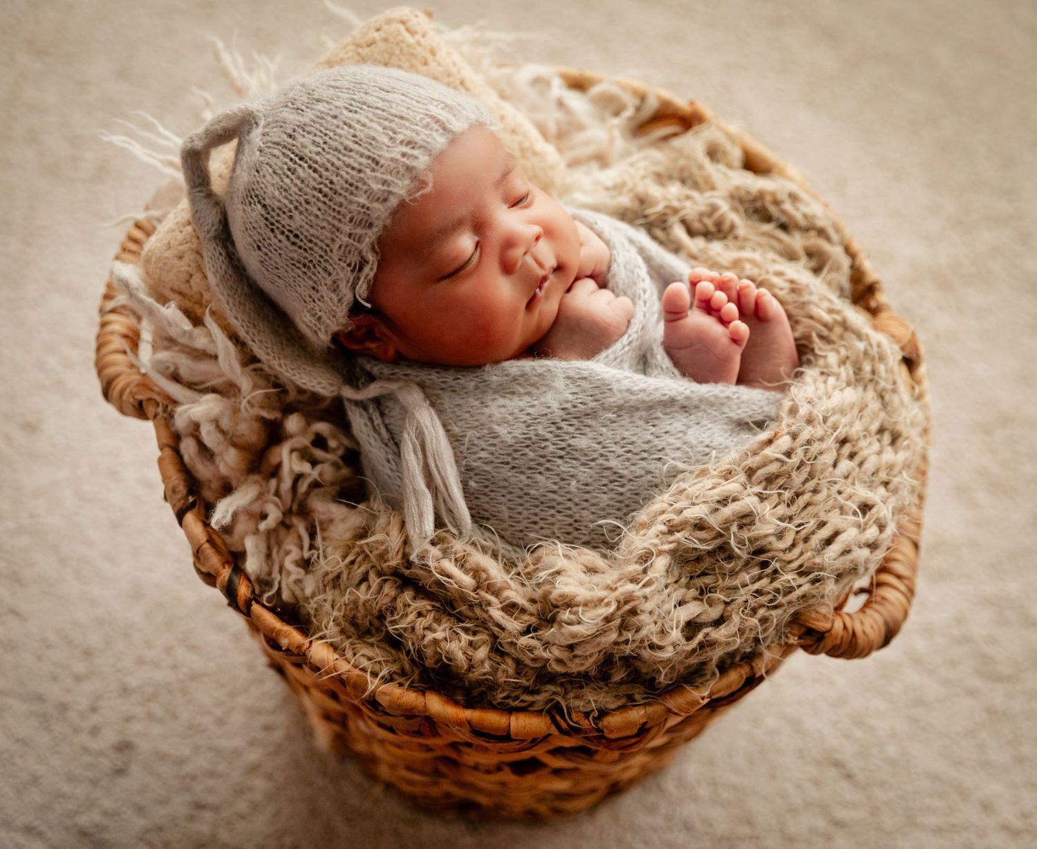 Newborn baby sleeping peacefully in a wicker basket, wrapped in a gray knit swaddle and matching bonnet.