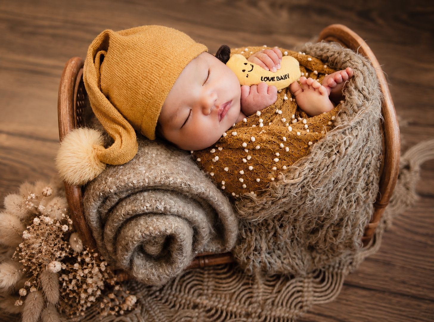 Sleeping newborn baby in mustard yellow outfit and bear hat resting in a wicker basket with soft blankets.